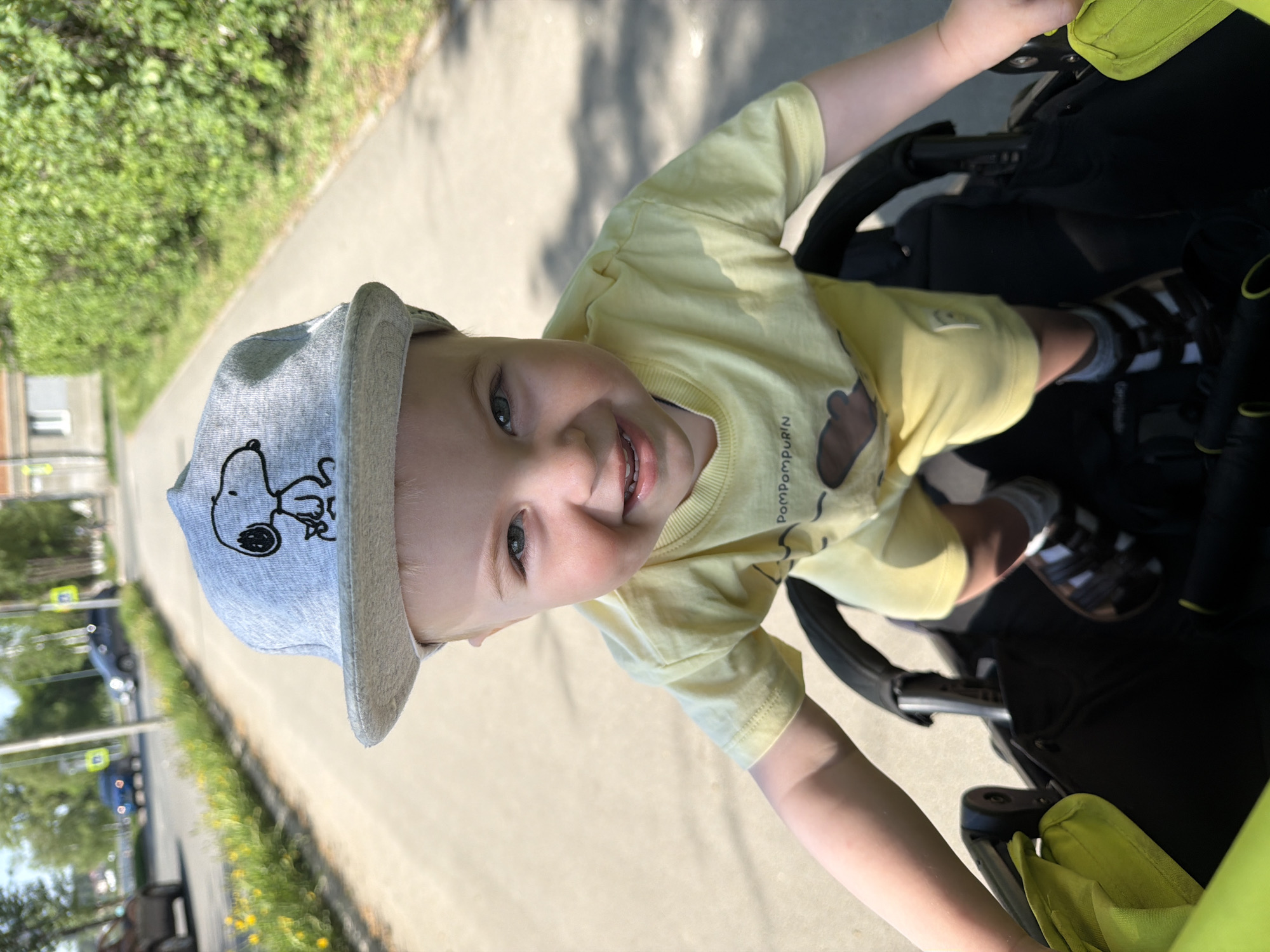 A child in a yellow t-shirt and a Snoopy cap sits in a stroller on a paved path, likely in a park. The path is bordered by greenery, and blurred vehicles suggest a nearby street. The location is Votkinsk, Russia.
