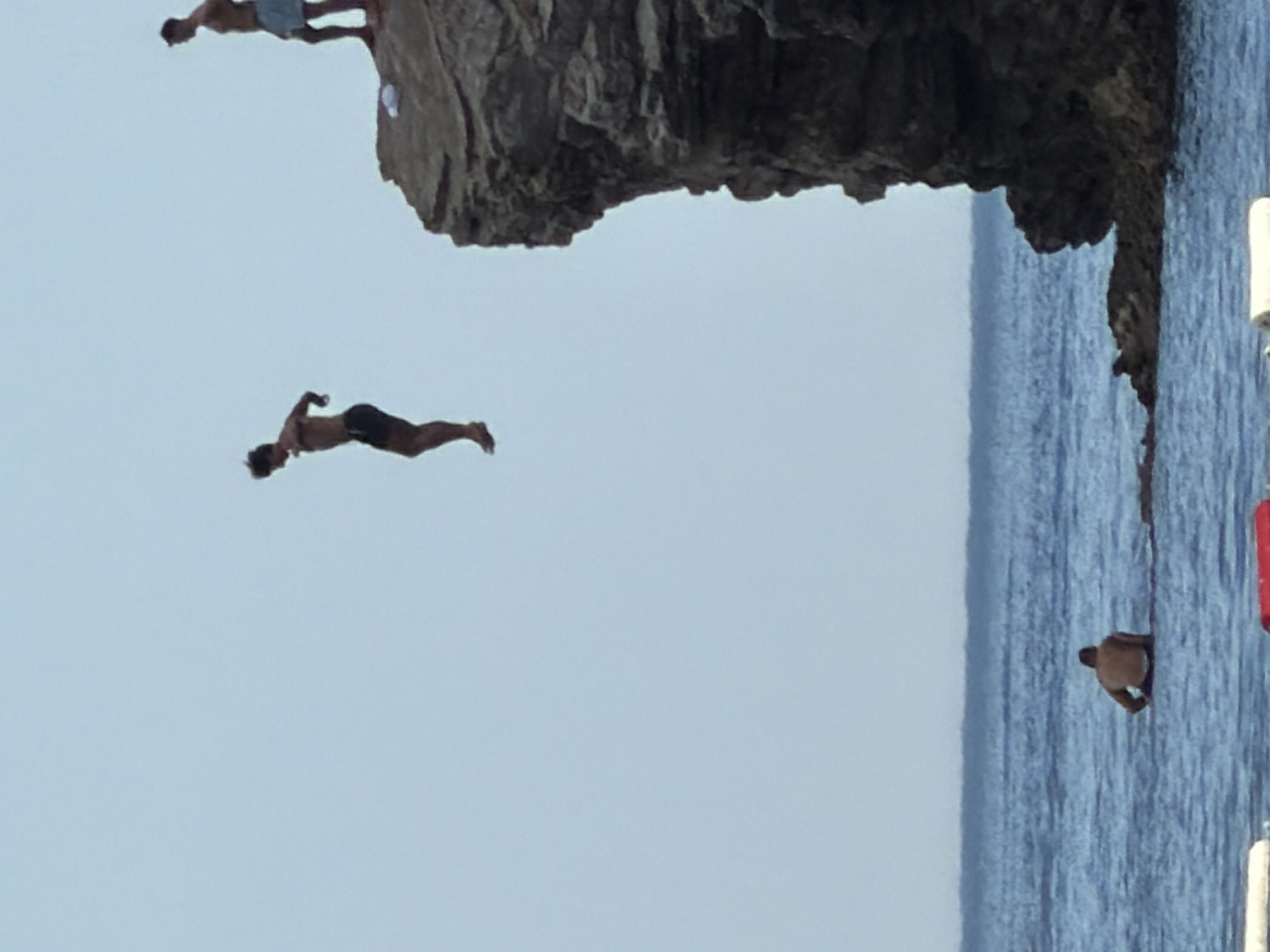 The image depicts a scenic coastal environment, likely in Budva, Montenegro, characterized by a clear sky and calm, blue water. In the foreground, the water is calm, with a person visible wading in it near the shore. In the mid-ground, a jagged rock formation juts out into the sea. On top of this rock, two individuals are present. One is standing and looking towards the sea, while the other is in mid-air, captured during a jump from the cliff. The jumper, a male, is dressed in dark shorts and is angled downwards as if in the process of plunging into the water. The overall scene conveys a sense of outdoor recreation and adventure.