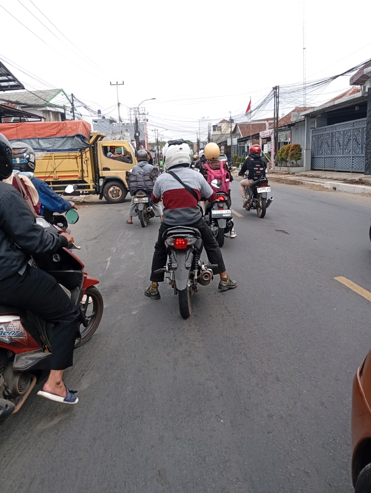 In a street scene in Tasikmalaya, Indonesia, several motorcyclists are navigating traffic. A yellow truck is visible on the left, its bed covered with an orange tarp. The road is asphalt, with some visible cracks and a yellow line marking the lane. Buildings line the right side of the street, with power lines crisscrossing overhead. Most of the individuals are wearing helmets for safety. One motorcyclist in the foreground is wearing black pants and a dark jacket. A rider behind them has a red and gray striped shirt, and another is wearing a pink backpack. The sky is overcast.
