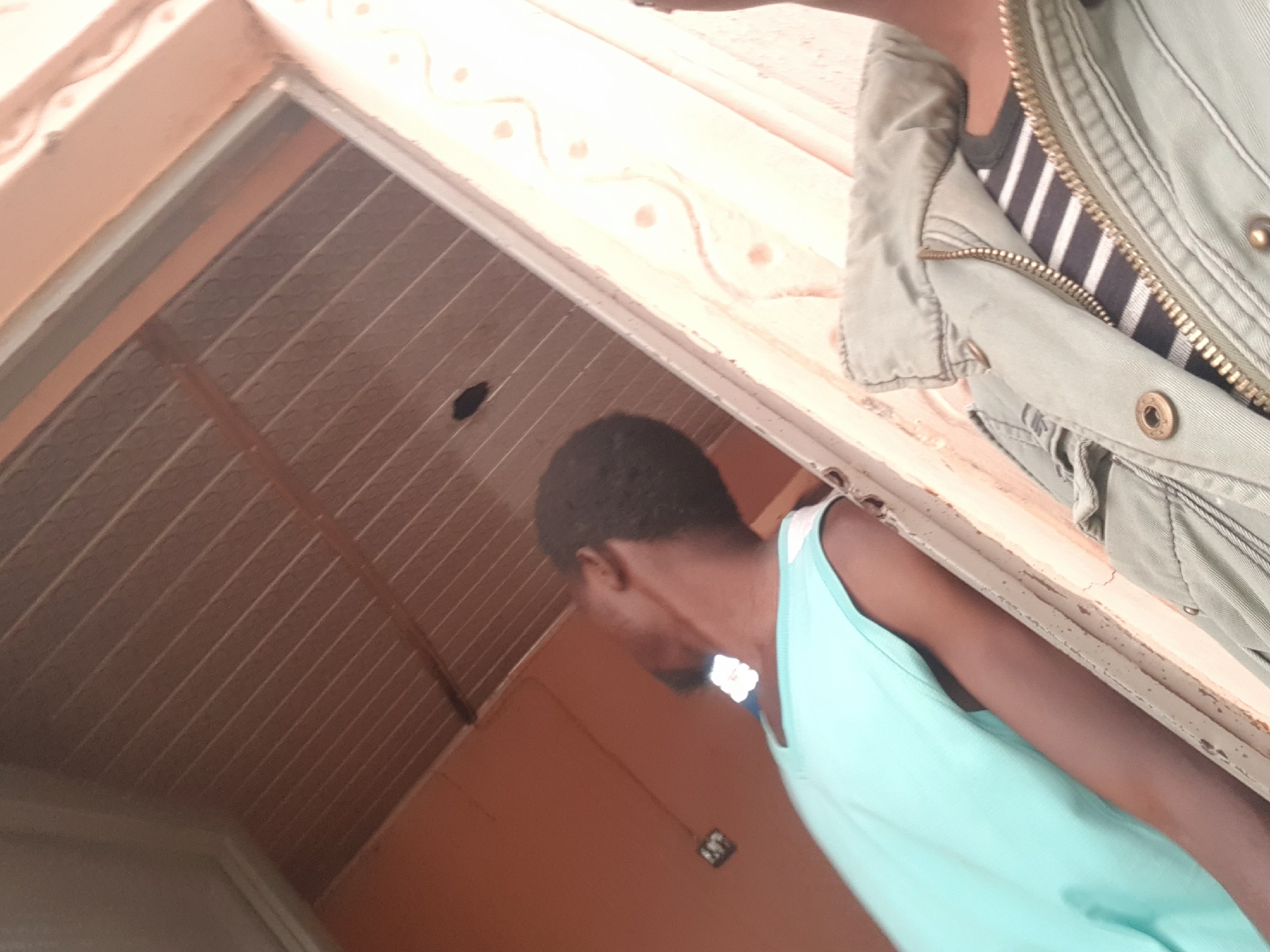 The image depicts a person in Dutsinma A, Nigeria, wearing a light blue tank top and partially obscured by a green jacket with a visible striped shirt underneath. They appear to be standing beneath a light-colored ceiling with recessed lines and a hole. A decorative border runs along the wall or frame. The walls are primarily orange-brown. The perspective is from a low angle, looking upwards at the subjects.