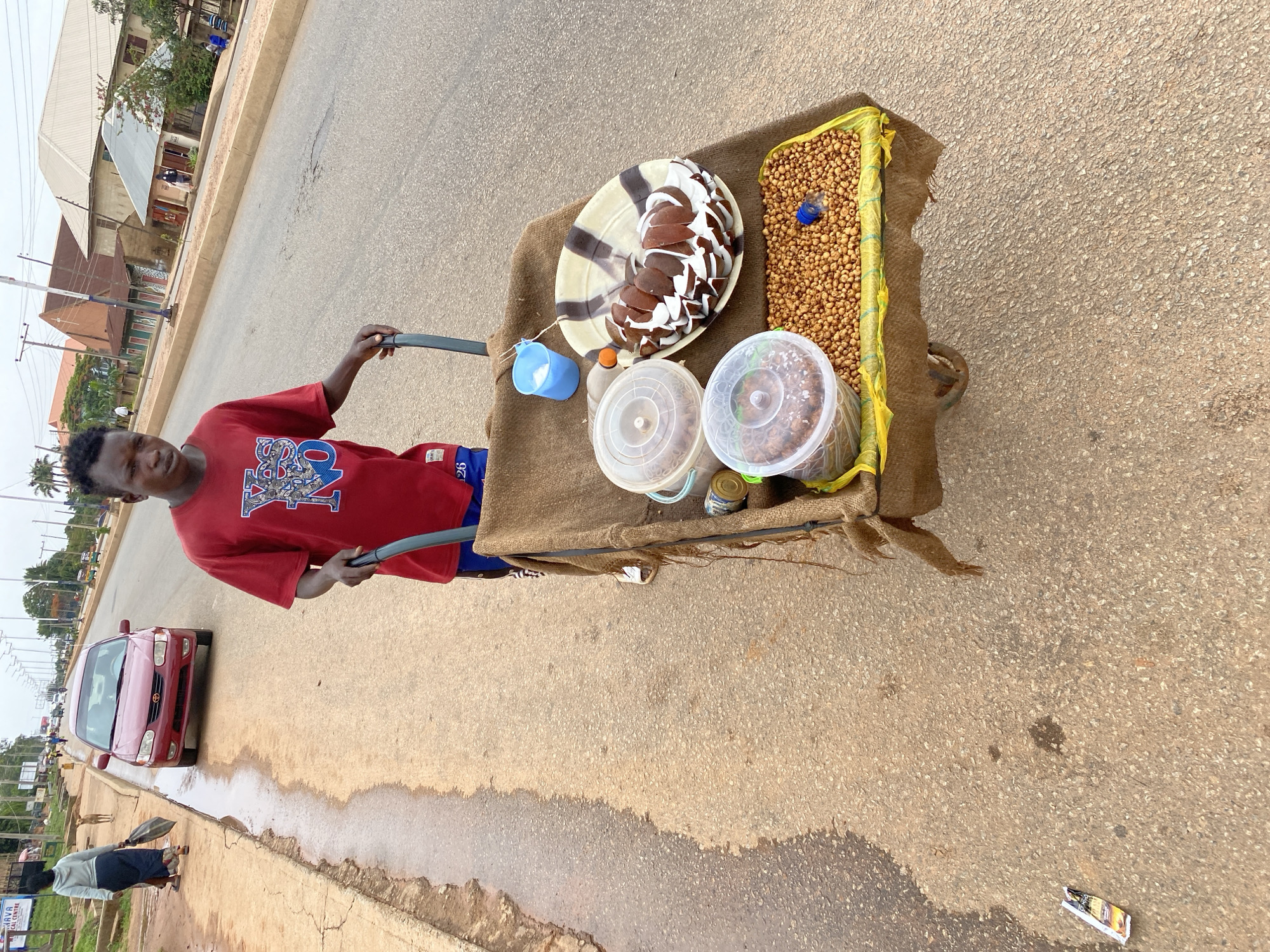 A street vendor is standing in the middle of a road in Bukuru, Nigeria, pushing a cart that is loaded with food items. The cart is covered with a brown burlap sack and contains a dish of sliced coconut topped with white frosting, two plastic containers of unidentified food items, a yellow bag filled with small round nuts, a small blue plastic cup, and other small containers. The vendor is a young, dark-skinned man wearing a red t-shirt and blue shorts. A red car is parked on the left side of the road, near the curb. Other people are walking along the sidewalk in the background. The road appears to be paved and has a curb on the left side. Buildings and trees are visible in the background.