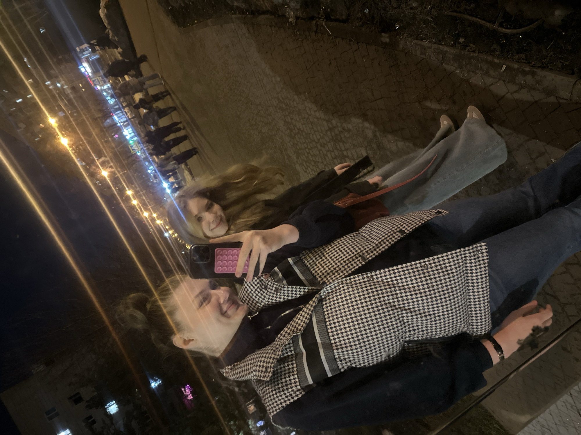 Two young women are taking a selfie using a mobile phone with a colorful case, reflected in a glass surface or a large mirror. One woman is wearing a black and white houndstooth-patterned jacket over a dark shirt and jeans, and has a wristwatch on her left wrist. The second woman is wearing a dark jacket with a red shoulder bag, light-colored jeans, and light-colored shoes. They are located outdoors in Kazan, Russia. In the background, there are buildings with illuminated windows and street lights, indicating it is nighttime. There are other people walking in the distance. The ground appears to be paved.