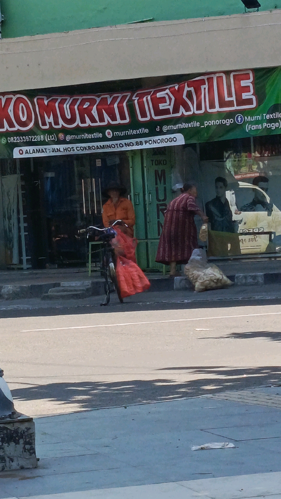 The image shows a street scene in Ponorogo, Indonesia, in front of a store called "KO MURNI TEXTILE". A banner with the store's name and contact information is displayed prominently. In front of the store, a woman wearing an orange shirt and a wide-brimmed hat stands next to a bicycle with a large red bag attached. Another woman in a striped dress stands further back, near a pile of goods. The street is paved, and a sidewalk runs along the front of the store. The overall lighting suggests it is daytime.