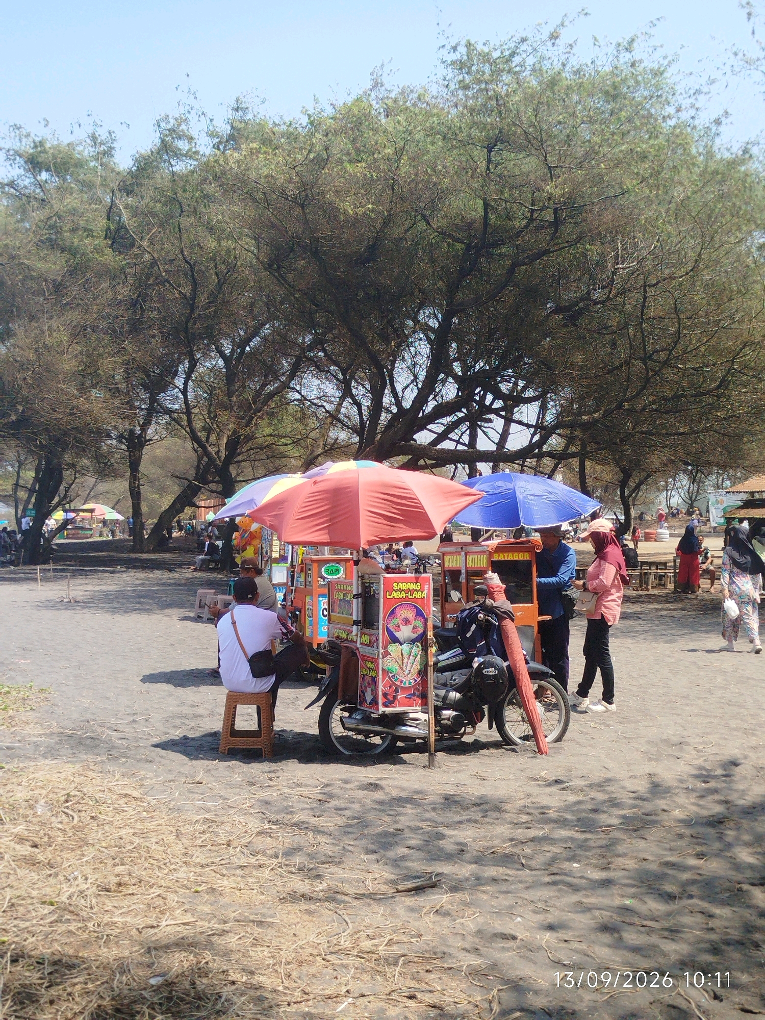 The image depicts a beach scene with several people and food carts. In the foreground, a person sits on a stool next to a food cart. The cart is adorned with colorful images and text. To the right, two individuals are standing near another food cart. One is interacting with the cart's vendor, and the other is looking on. In the background, several other food carts and people are visible, as well as trees providing shade. The ground is sandy, with some dry grass scattered around. The sky is clear and blue. The timestamp "13/09/2026 10:11" is visible in the lower right corner. The scene is likely a bustling beachside market in Pleret, Indonesia.