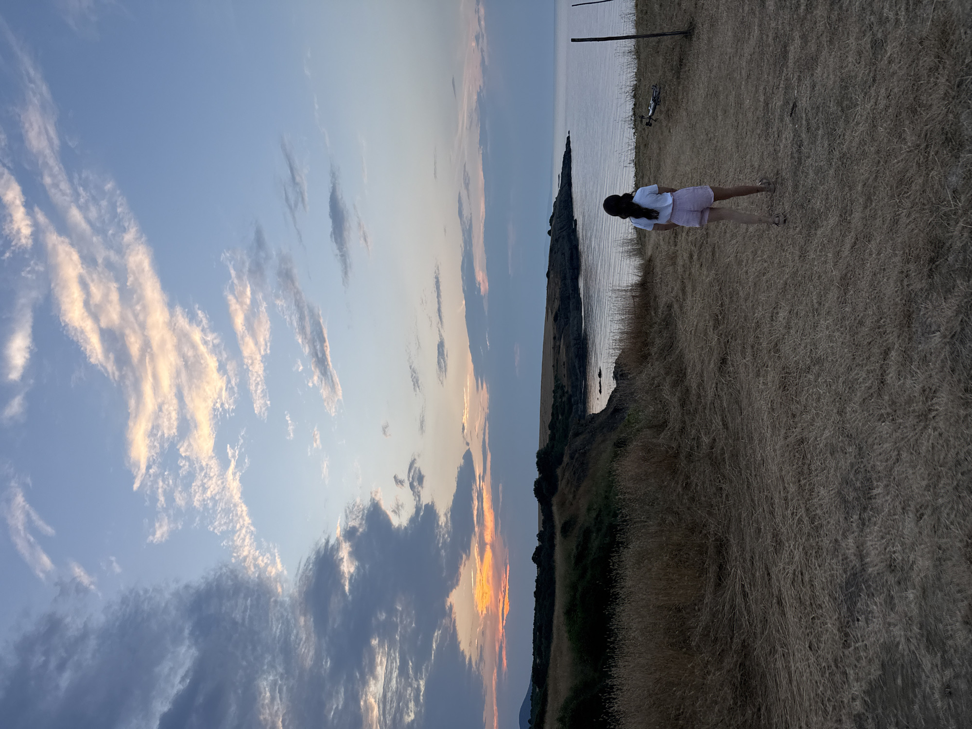 The image depicts a rear view of a young woman standing on a dry, grassy cliff overlooking the sea in Varvara, Bulgaria. The sky is a mix of blue and pale orange, with scattered clouds tinged with pink and yellow from the setting sun. The sea stretches out to the horizon, calm and reflecting the sky's colors. In the distance, a dark, tree-lined coastline curves to the left. The foreground is dominated by dry, straw-like grass. To the right of the woman, a dark pole and a collapsed drone are visible on the ground.