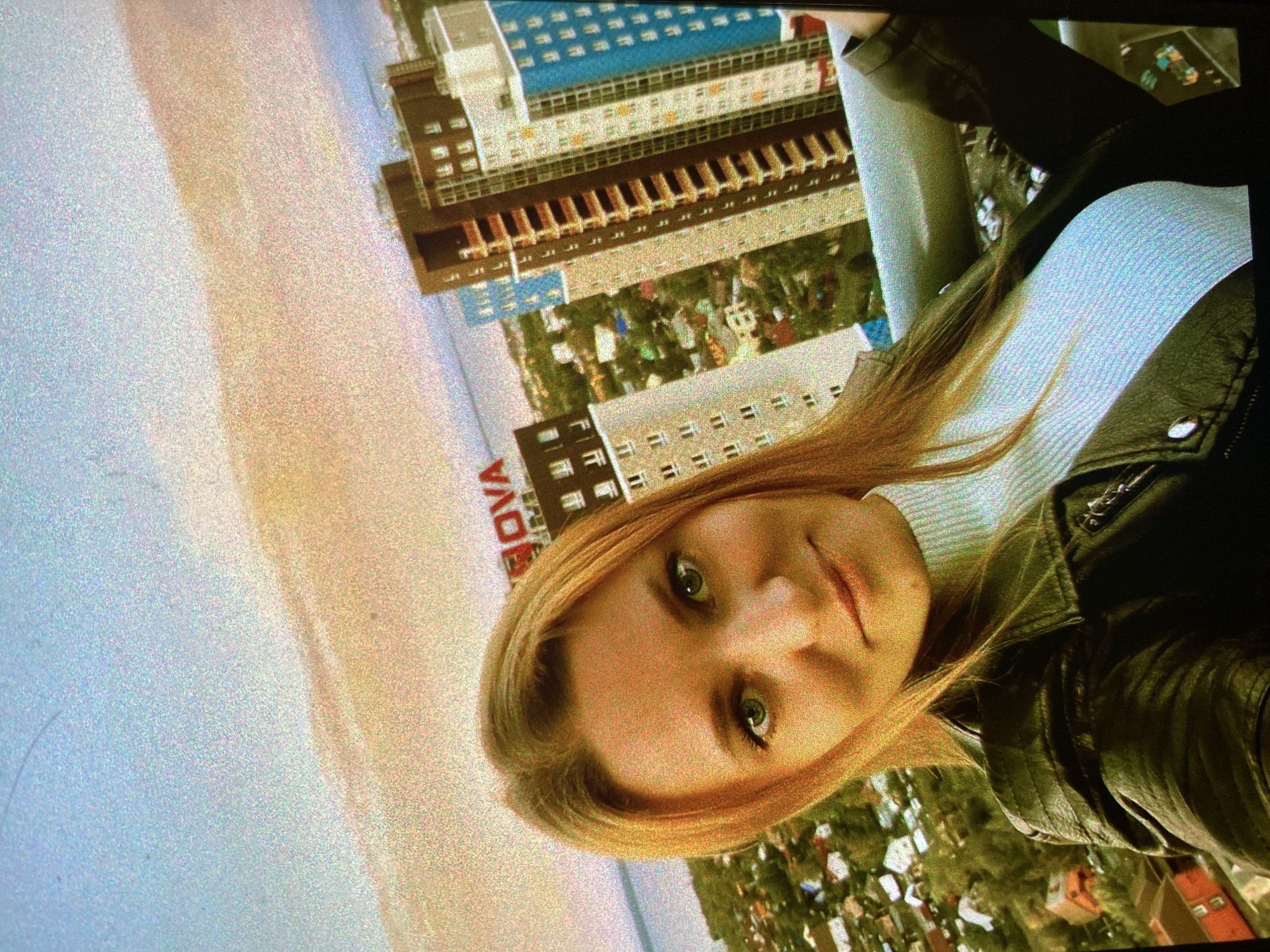 A grainy, rotated image depicts a young woman taking a selfie against an urban backdrop, likely in Saint Petersburg, Russia. The image is oriented with the woman's head on the right side of the frame.
The central subject is a woman with light brown or blonde hair and striking green eyes, looking directly at the camera with a neutral to slightly smiling expression. She is wearing a light-colored, possibly white or light blue, high-neck or turtleneck top under a dark leather jacket. Her hair falls loosely around her face and shoulders.
The background reveals a densely built-up urban environment with multiple multi-story buildings under what appears to be a daytime sky. One prominent building in the upper portion of the frame (as viewed in its current rotation) features a distinctive blue roof. Other buildings display various architectural styles, including one with prominent dark brown and light beige sections. Patches of green foliage and trees are visible between the structures. Towards the left side of the frame, a lighter, possibly sandy or paved area extends towards a bright, pale blue horizon, suggesting either a sky or a large body of water.
A notable detail is a partially visible red text "OVA" on a sign or building in the mid-ground, suggesting it could be part of a larger word or name. The overall image exhibits significant digital noise and graininess, giving it a somewhat retro or low-resolution appearance.
