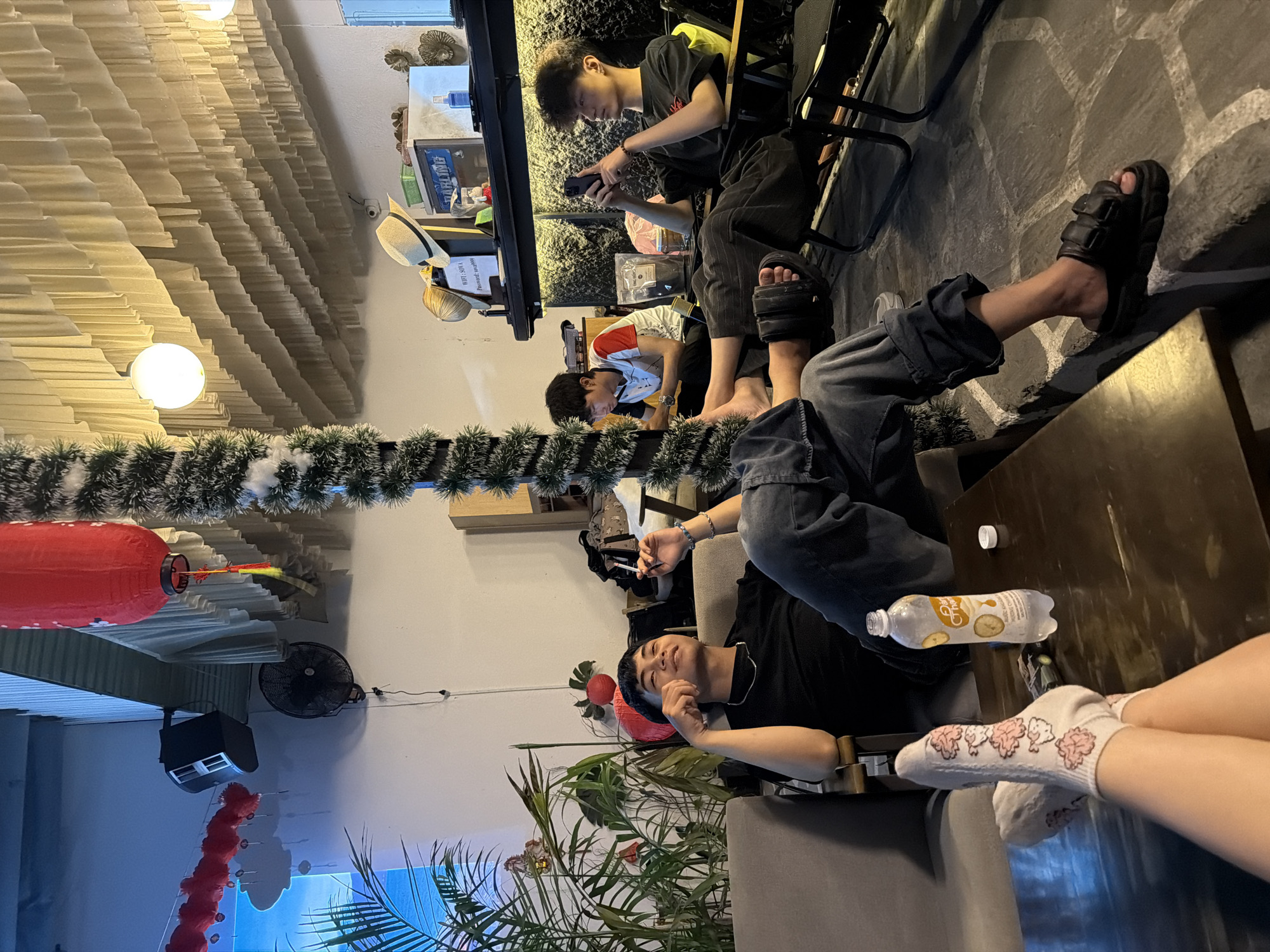 The image captures an indoor scene in Đà Nẵng, Vietnam. The primary focus is on several individuals relaxing in a café or similar establishment. A person wearing jeans and black sandals is reclining on a grey couch, with feet visible near a wooden table where a bottle of beverage is placed. Next to this person are legs with feet wearing socks patterned with what appears to be cartoon mushrooms. Another individual is seated, focused on a mobile device, dressed in a black t-shirt. Further in the background, there's another person looking at a phone in the same setting. The environment includes indoor plants, holiday garland, a ceiling fan, and decorative Chinese lanterns. The establishment's interior showcases a layered ceiling design. The lighting is ambient.