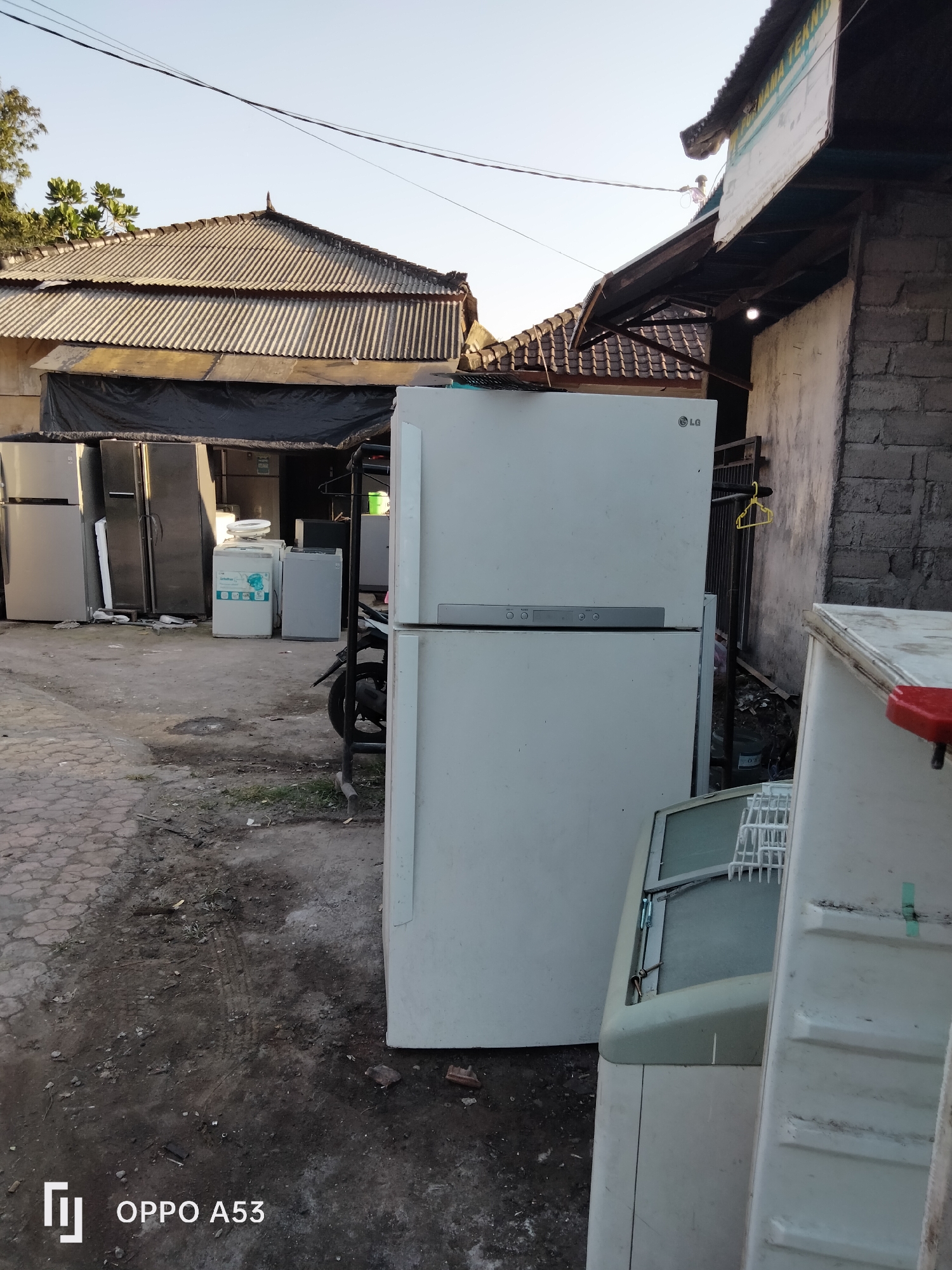 The image depicts an outdoor scene in Denpasar, Indonesia, likely an appliance repair or recycling yard. Dominating the foreground is a large white LG refrigerator, positioned in front of a weathered brick wall. To the right, another white appliance, possibly a chest freezer, is partially visible. In the midground, a row of various refrigerators and washing machines are lined up, some covered by a black tarp. A motorbike is parked behind the appliances. The ground is a mix of cracked paving stones and dirt. Overhead, power lines crisscross the sky, and a corrugated metal roof of a building is visible in the background, along with some foliage. The lighting suggests it is daytime, possibly late afternoon, with the sun casting shadows.