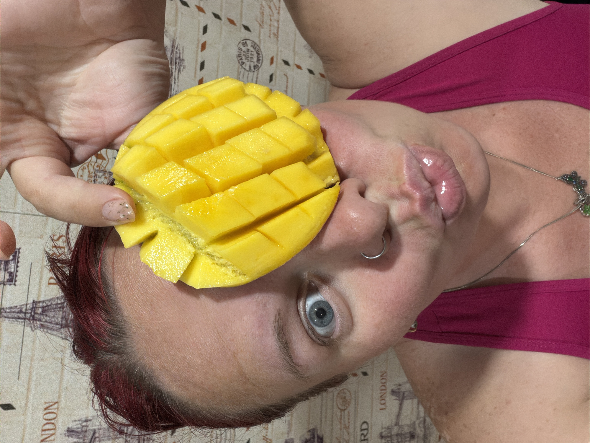 A woman with red hair and a nose ring holds a sliced mango to her face. Her eyes are blue. She wears a pink top and a silver necklace. The background shows a pattern with images of the Eiffel Tower and the words "Paris" and "London." There is no information in the image or its crops to indicate its connection to Chaikovsky, Russia.