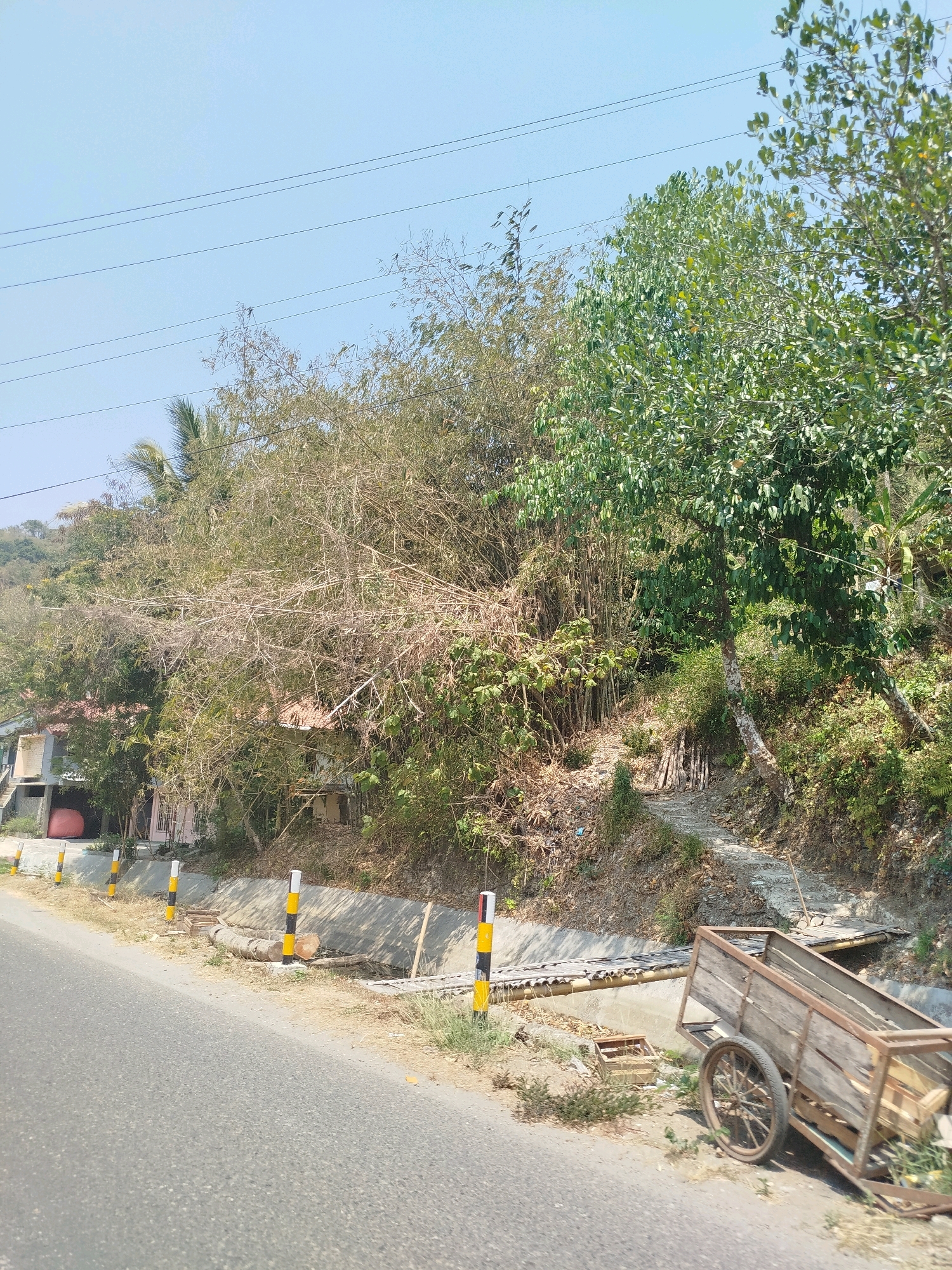 A rural road runs parallel to a drainage ditch on the left side of the image. Several black and yellow striped posts mark the edge of the road. A wooden cart with two large wheels sits on the right side of the road near the ditch. Beyond the ditch, there is a steep embankment covered in dense vegetation, including bamboo and trees. A path made of concrete stairs winds its way up the embankment. In the background, several small buildings are visible, nestled among the trees. The sky is clear and blue. The scene appears to be located in the city of Kutosari, Indonesia, based on the context.