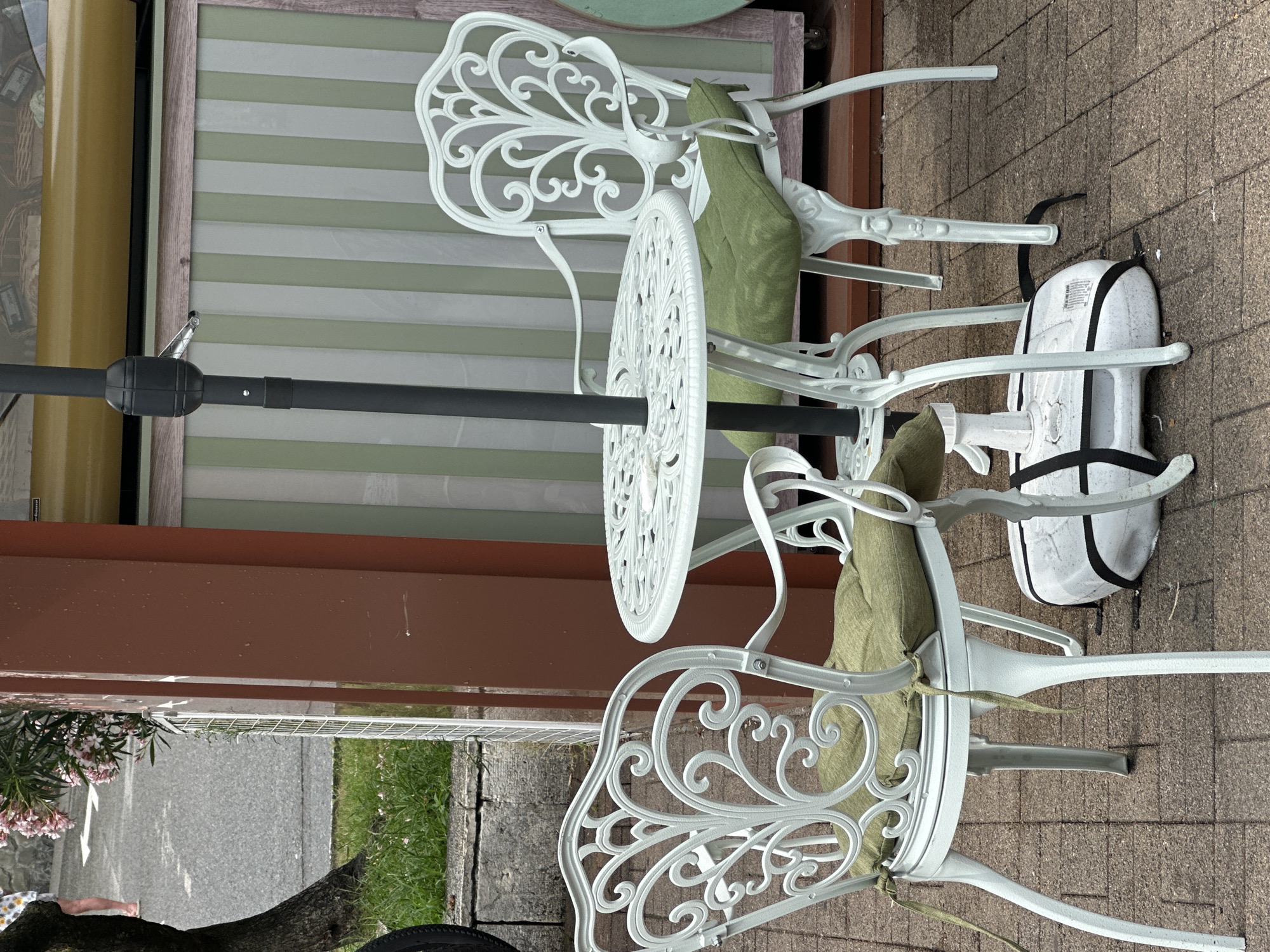 The image shows an outdoor seating area with a white cast-iron table and two chairs, each with green cushions. The table has an umbrella base in the foreground. The seating area is paved with brick. In the background, there is a building with green and white striped siding, and a reddish-brown column. To the left of the seating area, there is a tree trunk, grass, and a street with a person's legs visible walking. The scene appears to be in a cafe or restaurant setting. This scene is likely located in Sochi, Russia, given the context of the query.