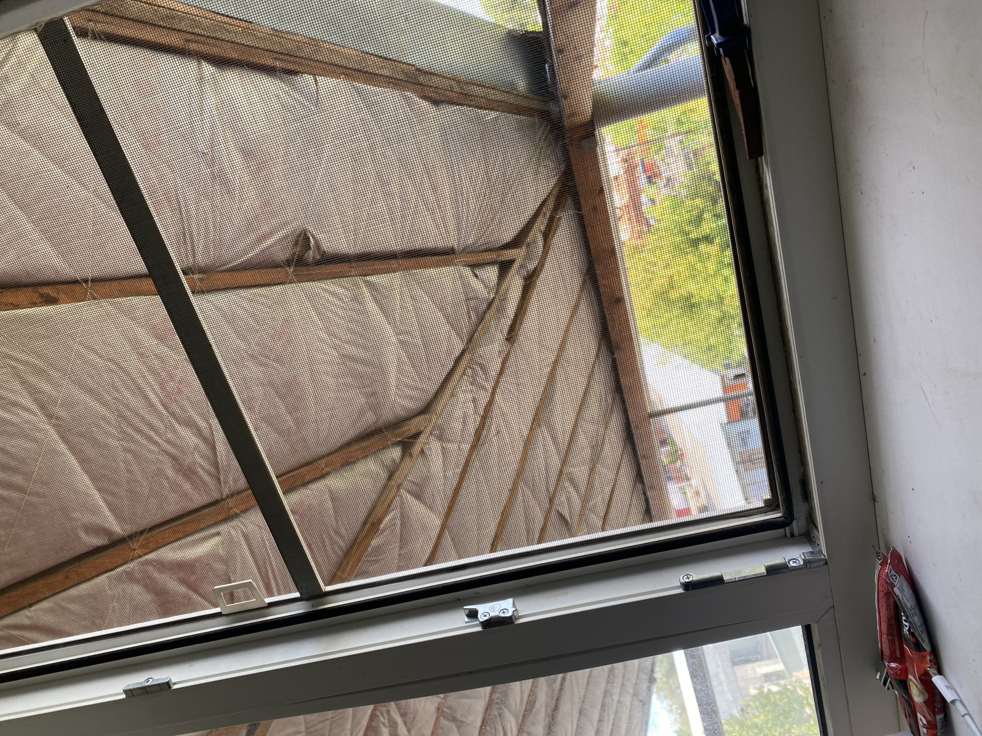 A close-up, angled interior view of a partially open window and its screen reveals a construction scene beyond. The window frame, made of white vinyl, is visible on the left, with a silver hinge attached to the left side. The screen, made of fine mesh, is partially open, allowing a view of a wooden roof structure with visible insulation. Outside, through the screen, glimpses of green trees and buildings can be seen. The windowsill in the foreground is white and shows some dust. On the windowsill, near the bottom left corner, are two red and silver foil-wrapped candy bars and a white cable. To the right, on the windowsill, is a piece of brown wood and a dark blue tool. The overall impression is of an unfinished construction or renovation project. The location is identified as Dedovsk, Russia.