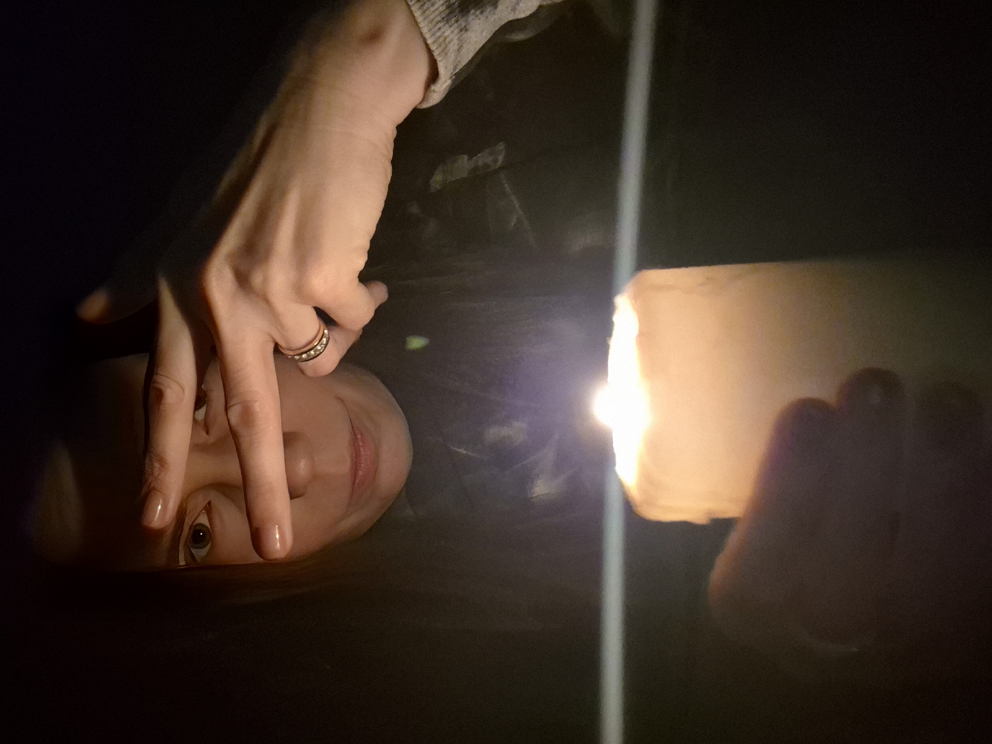 The image presents a close-up, low-light scene featuring a person's partially visible face and hand, illuminated by a bright artificial light source, likely a flashlight. The photograph is oriented vertically, with the person's head positioned on the left side, looking towards the right where the light source is intensely bright.
The individual's skin is pale, with parts of their face, including one eye, a portion of the nose, and lips, visible and directly lit. The visible eye is open and appears to be gazing towards the light, while a subtle smile is present on their lips. A hand, adorned with a gold-toned ring featuring several small, embedded stones on the ring finger, is positioned across the upper part of the face, with fingers resting over the forehead and brow area. The person's arm is visible with a light-colored, possibly knitted, sleeve.
The background is almost entirely dark, indicating that the scene is set in a very dimly lit environment, possibly indoors at night. The powerful beam from the flashlight creates a stark vertical streak of light on the right side of the frame, strongly illuminating the light source itself and casting highlights on the person's features. The activity suggests an interaction with the light source, perhaps inspecting something or simply using the light in a dark space, with the hand potentially shielding the eyes from the brightness or resting contemplatively. There is no visible text in the image. The scene is located in Chaikovsky, Russia.