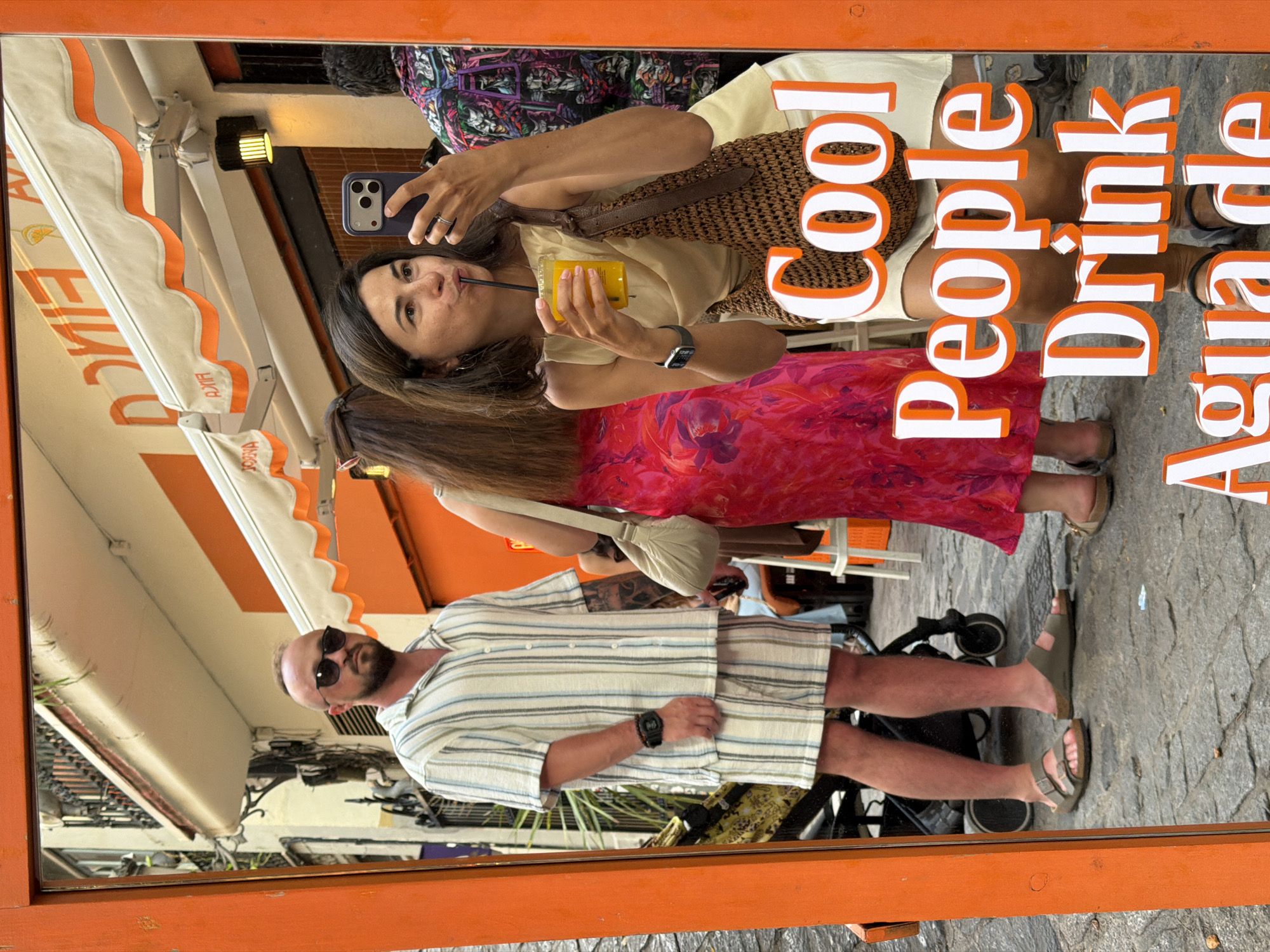 A woman in a tan top and white skirt takes a selfie in a mirror, holding an orange drink and a phone. A man in a striped shirt and shorts stands to her left, and another person in a pink dress is partially visible to her right. Text overlays the image reading "Cool People Drink Agua de". The setting appears to be a street in a city, with buildings and awnings visible in the reflection.