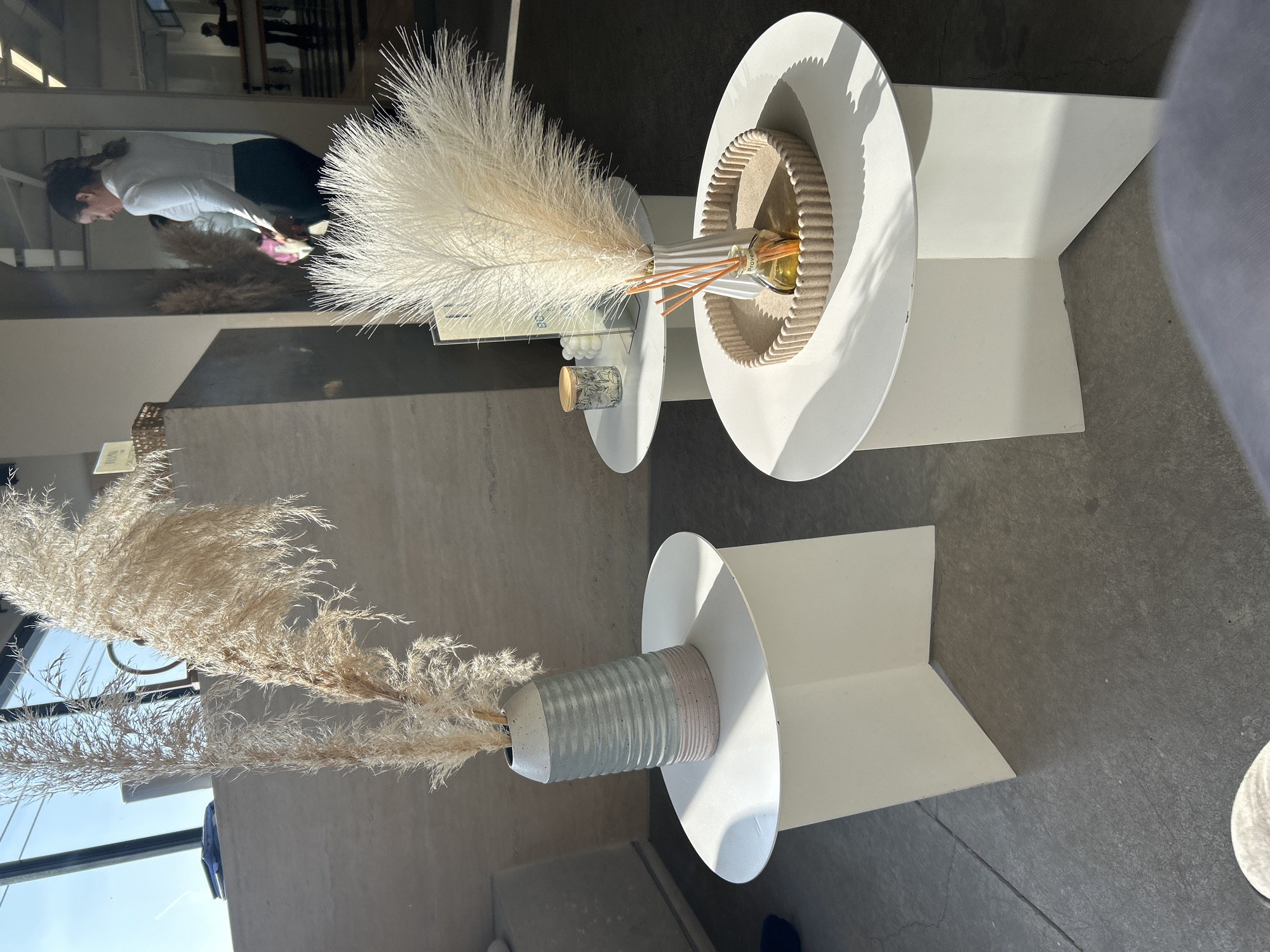 The image shows two vases with fluffy, wheat-colored stems, placed on white, modern pedestals with round tops and angular bases. The location appears to be indoors, possibly a store or showroom, based on the interior design and the person reflected in the window, who seems to be browsing. The setting gives the impression of a carefully curated display.