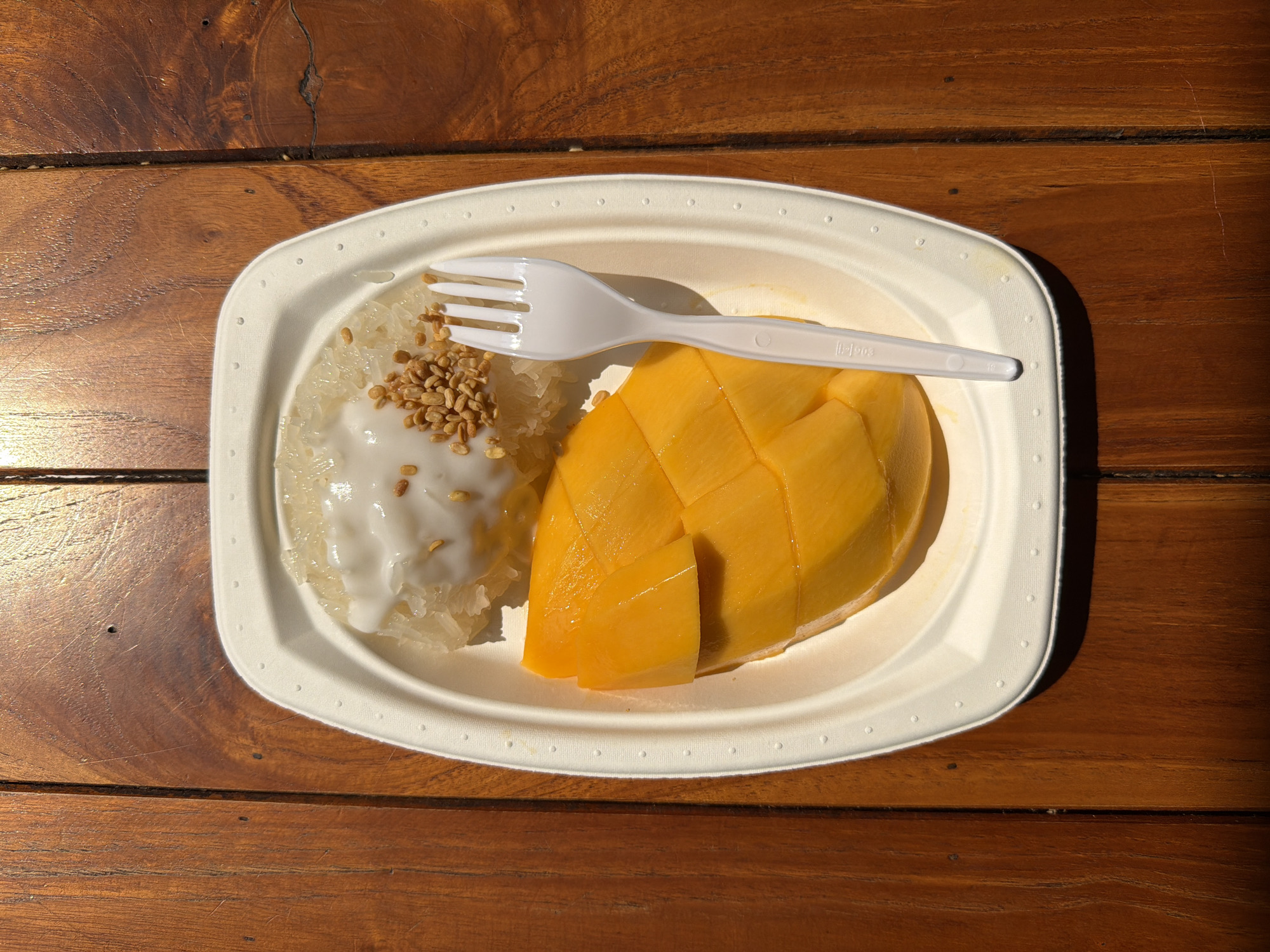 On a wooden table in Bangkok, Thailand, sits a white rectangular plate holding mango sticky rice. The mango, sliced in a grid pattern, is positioned on the right side of the plate, while the sticky rice, topped with a white sauce and sesame seeds, is on the left. A white plastic fork rests diagonally across the dish. The lighting suggests an outdoor setting.