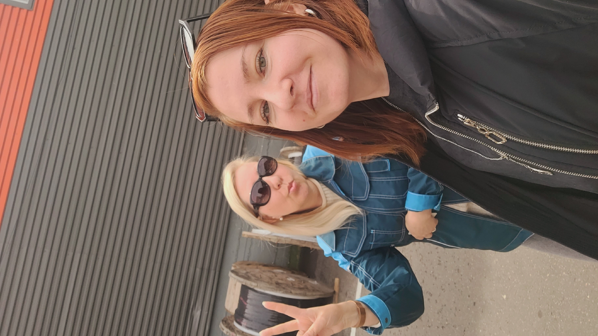 Two women pose for a selfie outdoors. The woman in the foreground has reddish-brown hair and is wearing glasses pushed atop her head and a black jacket. Behind her to the left, the other woman has blonde hair, sunglasses, and is wearing a blue jacket with turquoise accents. She is holding up two fingers in a peace sign. Behind them is a gray corrugated metal wall and to the left is a red stripe. In the foreground, there are two spools of black wire. The location is in the city of Unknown, Unknown.