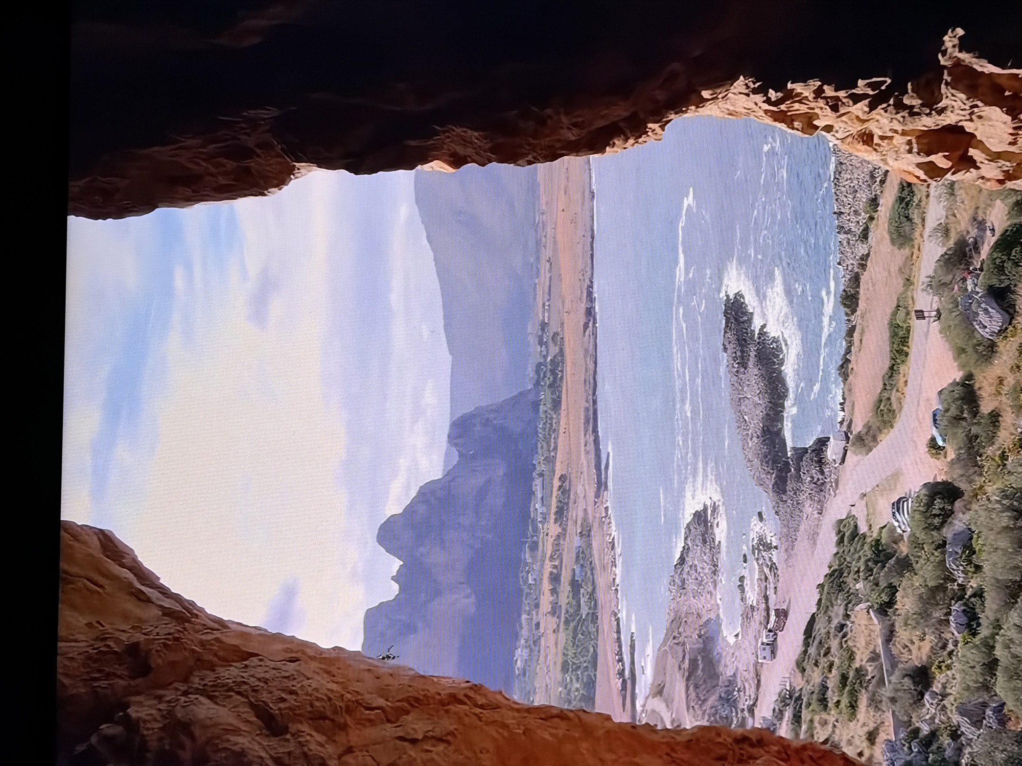 The image shows a view from a cave opening, looking out over a coastal landscape. In the foreground, the rough, reddish-orange rock of the cave walls frames the scene. Beyond the cave, a body of water, likely the sea, meets a rocky coastline. Several cars and what appear to be recreational vehicles are parked along a road that follows the coastline. In the distance, mountains rise from a dry, undulating landscape dotted with sparse vegetation and some buildings. The sky is partly cloudy.
This scene is NOT located in Chirchik, Uzbekistan. Chirchik is an inland city in Uzbekistan and does not have a coastline or coastal mountains like those depicted. The landscape suggests a Mediterranean or similar coastal environment.