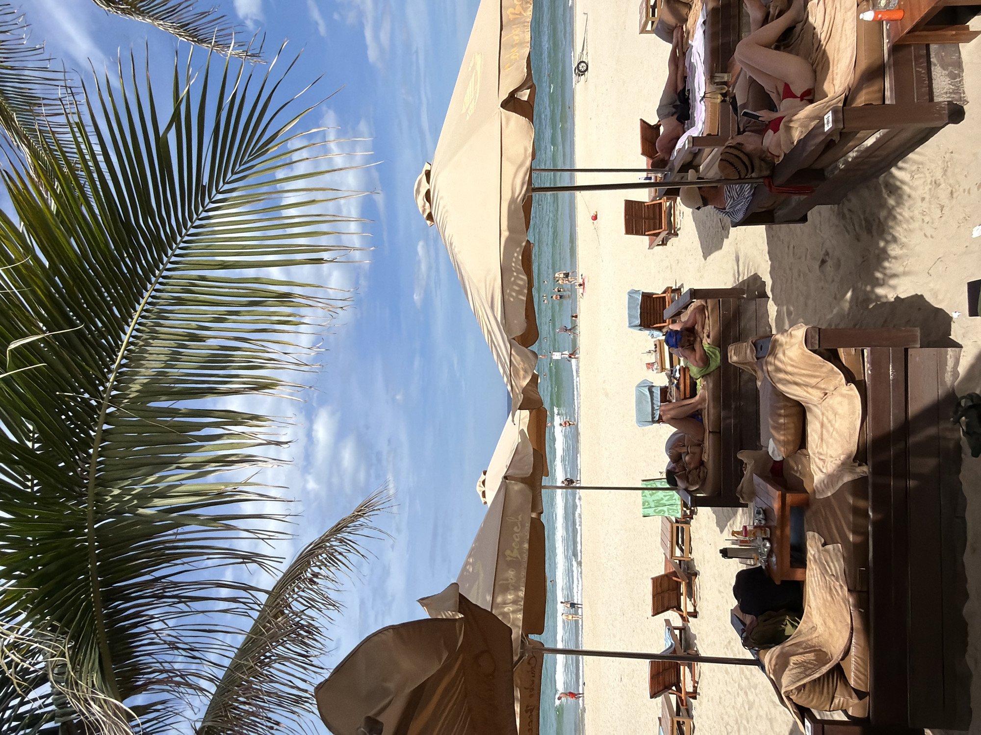 The image depicts a beach scene at My Khe Beach in Da Nang, Vietnam. In the foreground, several wooden lounge beds covered with tan cushions and blankets are arranged on the sand, some occupied by individuals. One person is lying down, while another is seated, looking at their phone. To the left, a small wooden table holds drinks and a container. Behind these lounge areas, beige beach umbrellas provide shade. The middle ground shows the sandy beach with more lounge chairs and umbrellas. A few people are walking on the beach or near the water's edge. The ocean is calm with gentle waves, and several people are visible in the water. The background features a clear blue sky with scattered white clouds. Palm fronds hang into the frame from the top, suggesting a tropical setting. The overall atmosphere is one of relaxation and leisure.