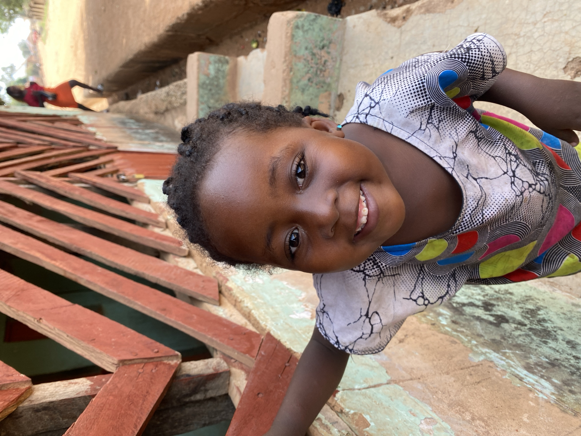 A young girl with dark skin and braided hair is smiling and looking directly at the camera. She is wearing a short-sleeved shirt with a busy white, black, and colored pattern. The background shows the environment of the city of Kuru Dungus, Nigeria, including wooden planks, possibly part of a structure or building under construction. An adult figure can be seen in the background, as well as a dilapidated wall.