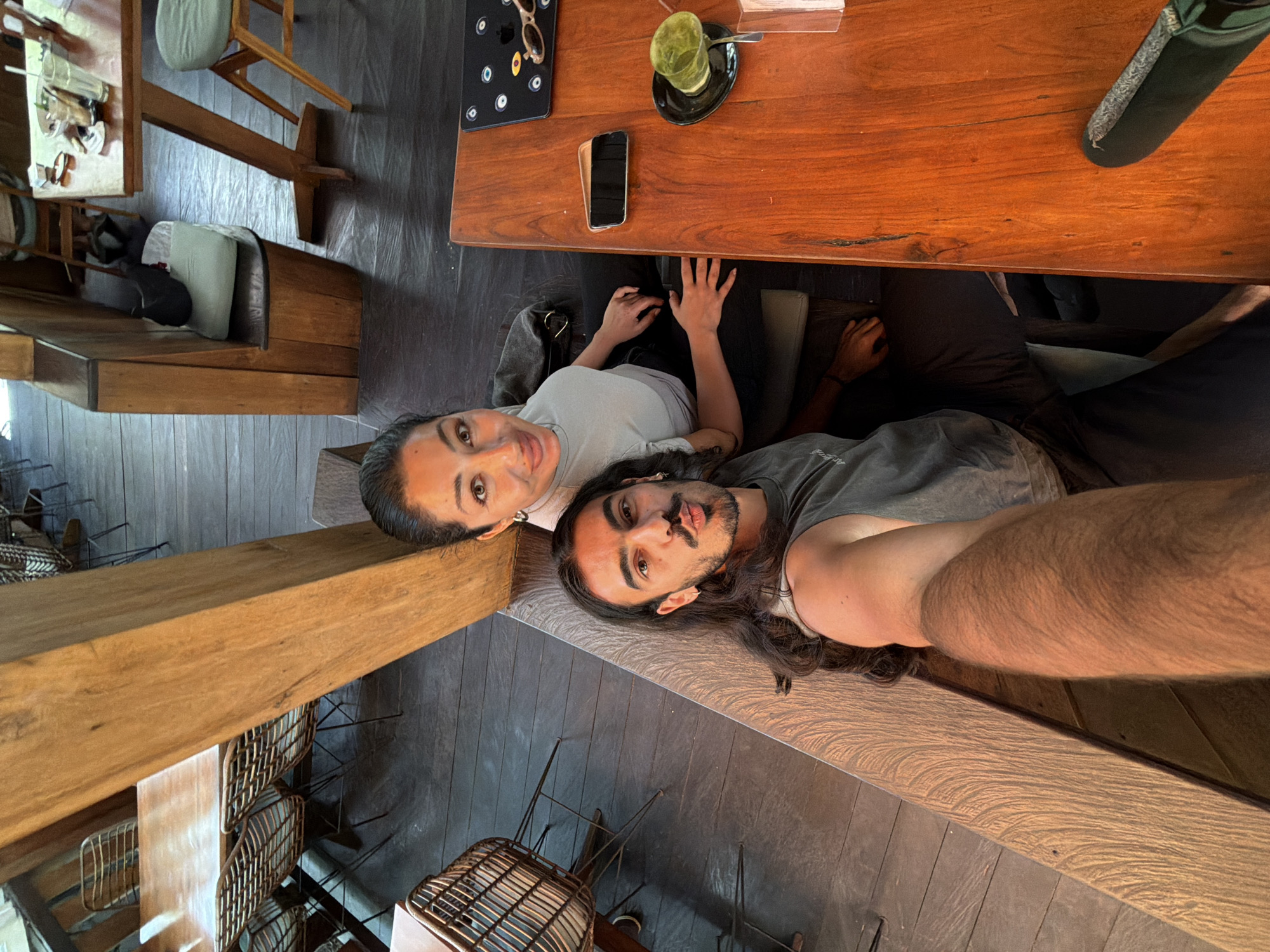 Two individuals, a man and a woman, are captured in a selfie-style photograph taken from a slightly elevated angle. They are seated together at a large wooden table in what appears to be a cafe or restaurant in Kuta Selatan, Indonesia.
The woman, on the left, wears a light-colored t-shirt and has dark hair pulled back. She looks directly at the camera with a soft, pleasant expression. The man, on the right, has long dark hair, a beard, and a mustache, wearing a dark grey t-shirt. He also looks towards the camera, with his right arm extended, suggesting he is holding the camera.
The sturdy wooden table separates the subjects from the upper portion of the image. On the table, several items are visible: a smartphone lying face down, a pair of sunglasses resting on a dark mat with circular patterns, and a glass containing a green drink (possibly matcha or a green juice) with a straw, placed on a dark saucer with a spoon. A small white container or box is also on the table.
The interior of the establishment features predominantly wooden furniture, including benches or booths where the subjects are seated, and other tables and chairs with wicker-like backs in the background. The floor is dark. The overall ambiance appears relaxed and casual, characteristic of a daytime cafe setting. No discernible text is visible in the image.