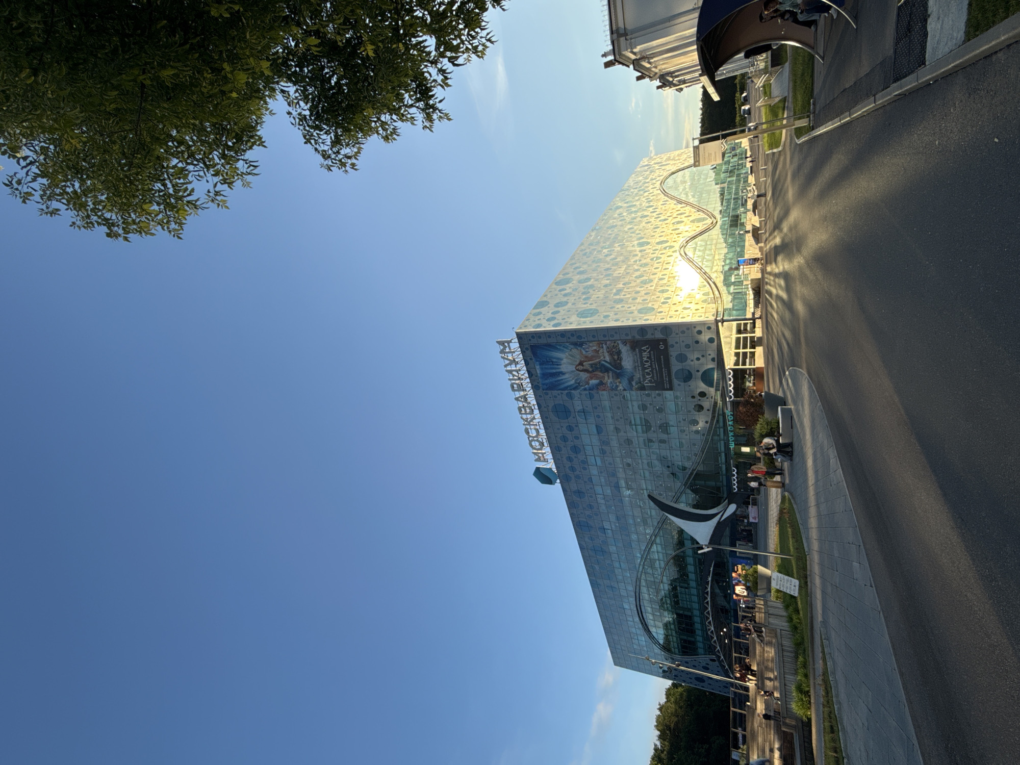 This image captures the exterior of the Mosquarium (Москвариум) building in Moscow, Russia, on a bright, sunny day. The scene is illuminated by strong, directional sunlight, casting long shadows on the paved road and sidewalk, suggesting either a late afternoon or early morning setting. The sky above is a clear, vibrant blue with only a few wispy clouds.
The central subject is the large, modern Mosquarium building, which boasts a distinctive glass facade adorned with a pattern of light blue circular elements and undulating, wave-like architectural features. The sun reflects brilliantly off its surface, creating shimmering light patterns. Large blue letters spelling "МОСКВАРИУМ" (MOSKVARIUM) are prominently displayed on the upper part of the building. Below this, a significant banner features an artistic depiction of a mermaid or mythical sea creature, with the Russian word "РУСАЛОЧКА" (RUSALOCHKA, meaning "Little Mermaid" or "Mermaid Show") clearly visible.
In the foreground, lush green foliage from trees partially frames the top left corner of the image. A paved road curves through the scene, accompanied by a wide concrete sidewalk. Scattered around the entrance area of the building and along the path are several individuals and small groups of people, some walking, others seemingly standing or sitting. A modern, abstract sculpture, dark in color, stands near the building's entrance. The overall impression is one of a busy, public attraction bathed in natural light.