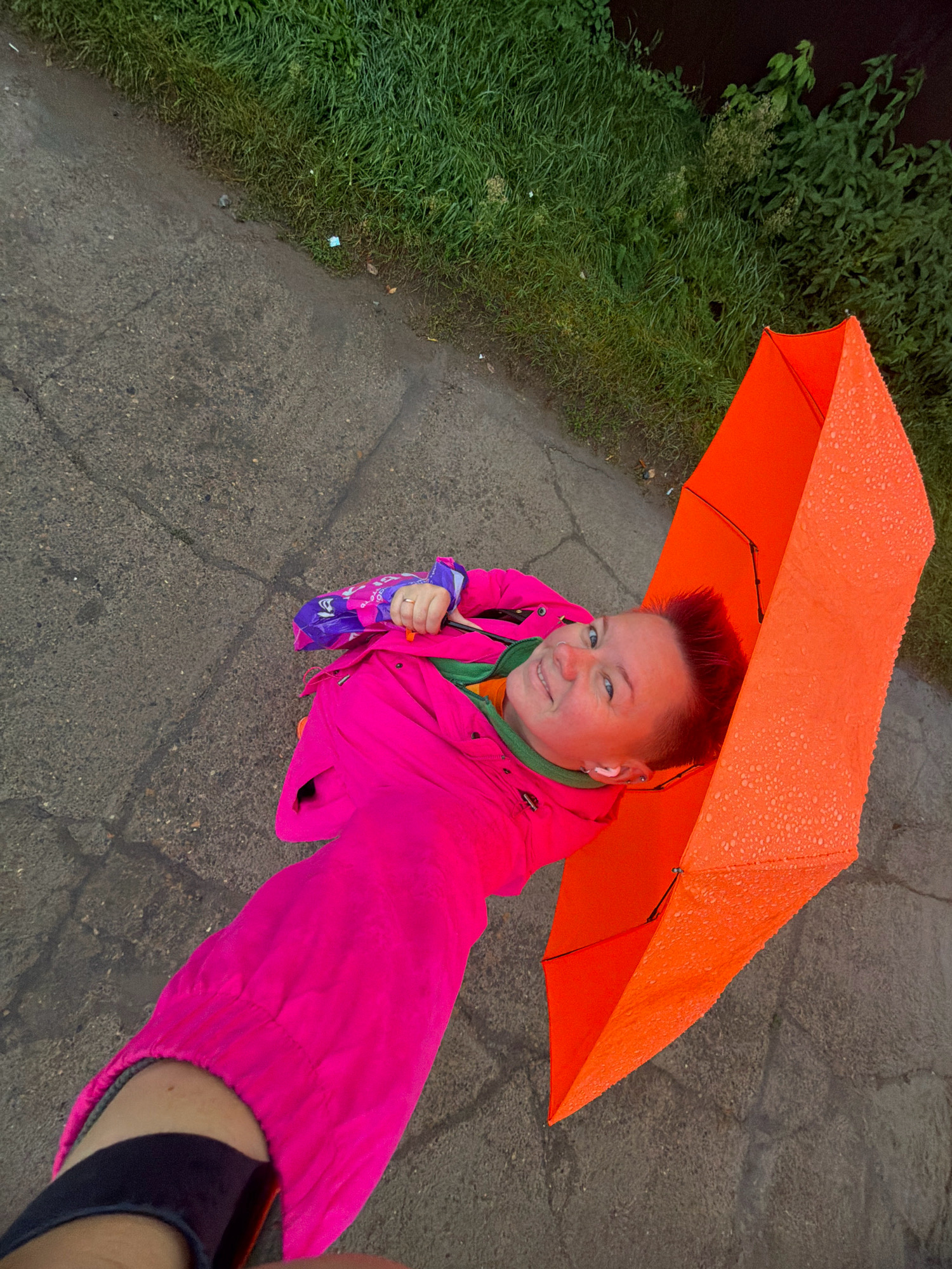 A person, wearing a bright pink hooded jacket and an orange umbrella, is pictured outdoors. The person's face, with short, spiky red hair and visible ear buds, is turned towards the camera, smiling. They are holding a purple and blue object in their left hand. The scene appears to be a street with a paved surface, bordered by a grassy area. Water droplets are visible on the umbrella, suggesting it may be raining or has recently rained. The image is taken from a high angle, looking down. The location is identified as a street in Chaikovsky, Russia.