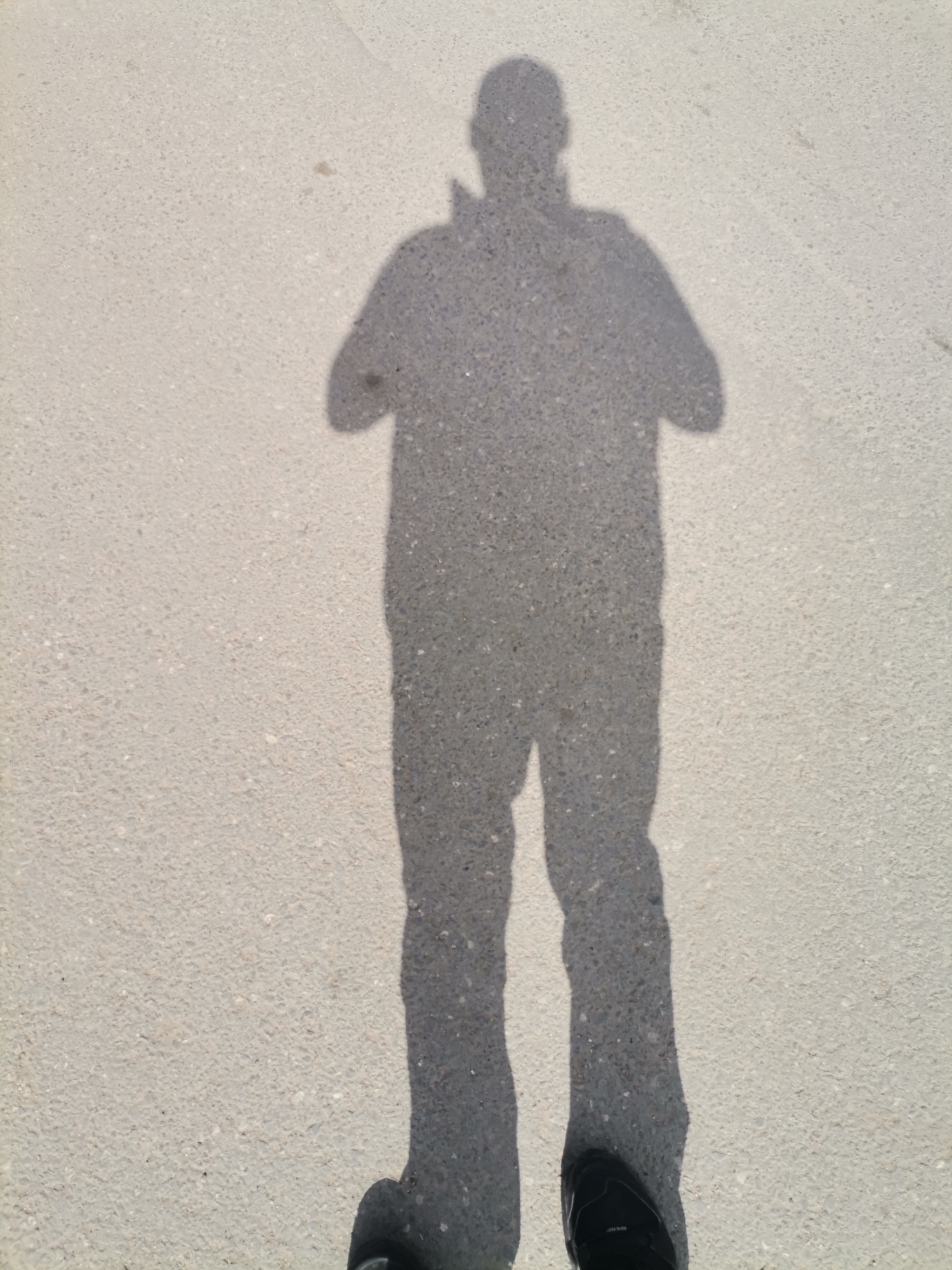 The image depicts the shadow of a person standing on a concrete surface. The shadow suggests the person is wearing pants and a collared shirt or jacket. The person's hands appear to be held in front of them. The concrete surface is light gray and textured. A portion of one shoe, presumably belonging to the person casting the shadow, is visible at the bottom of the image. The image does not provide information to determine the location is in the city of Dobrich, Bulgaria.