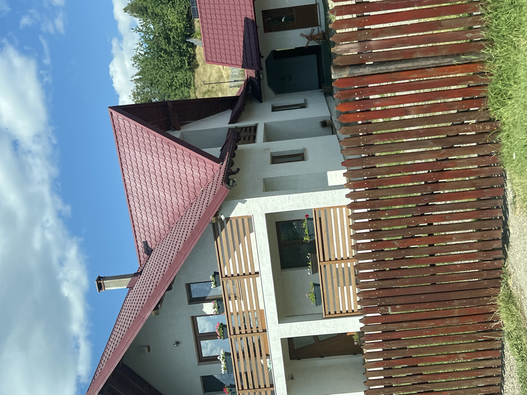 A detached house with a red metal roof and white walls is pictured in Capu Codrului, Romania. The house features multiple windows and balconies with wooden railings. A wooden fence runs along the front of the property. Behind the house, a forested hillside is visible under a bright blue sky with scattered clouds.