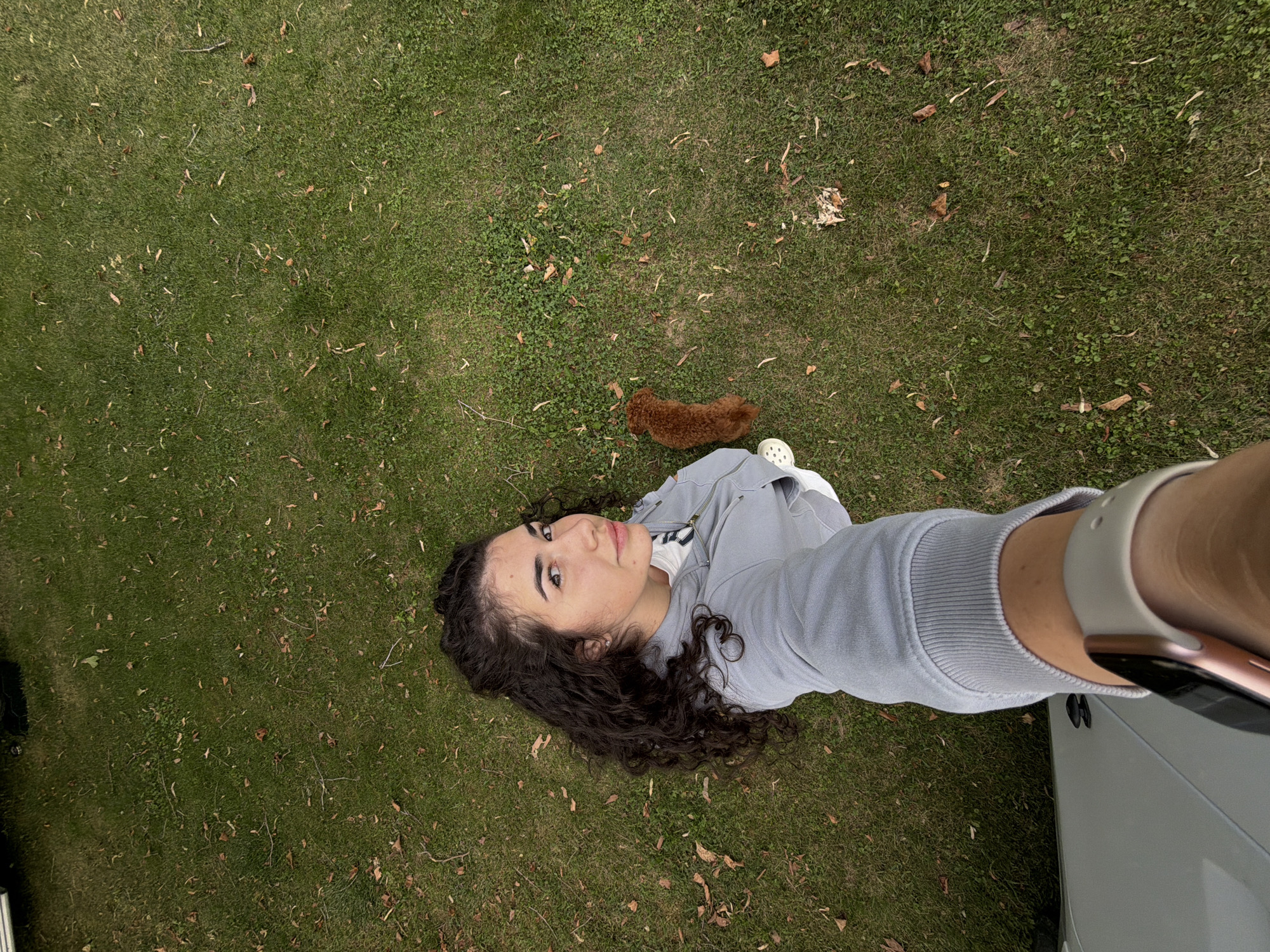An overhead, wide-angle selfie shows a woman with dark, curly hair standing on a patch of grass, with a small, reddish-brown dog to her right. She is wearing a light gray hooded sweatshirt, a white t-shirt, and light-colored sneakers. Her left arm is extended, holding the camera. The background is a lawn with scattered fallen leaves. The woman is looking at the camera. There is a partial view of a light-colored vehicle to the left of the frame. This image was taken in Kazanlak, Bulgaria.
