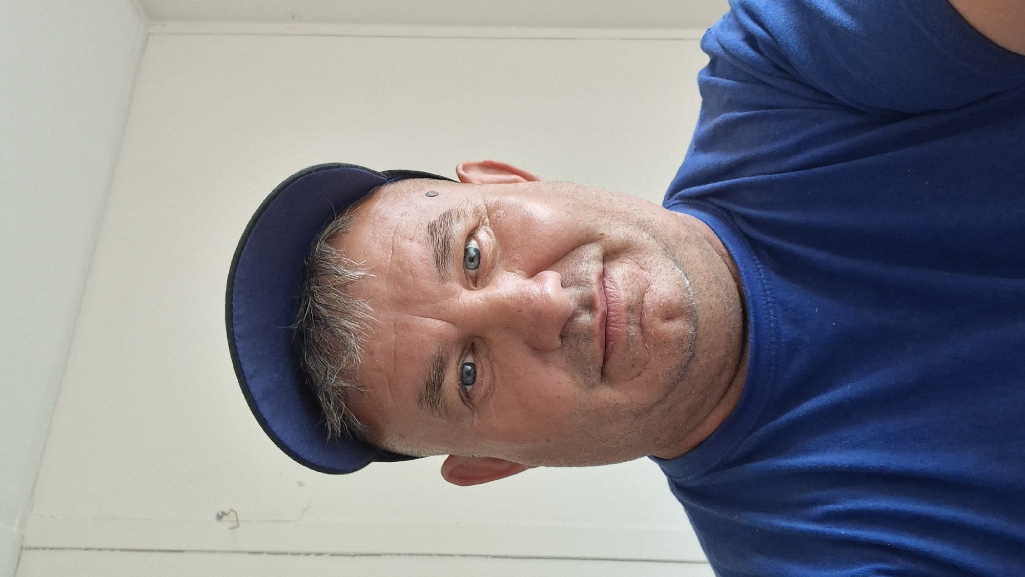 A man with short grey hair, wearing a blue t-shirt and a blue baseball cap, is pictured in a close-up portrait. He has blue eyes and a light complexion. He is outdoors, with a white wall behind him. The image was taken in the city of Hîncești, Moldova.