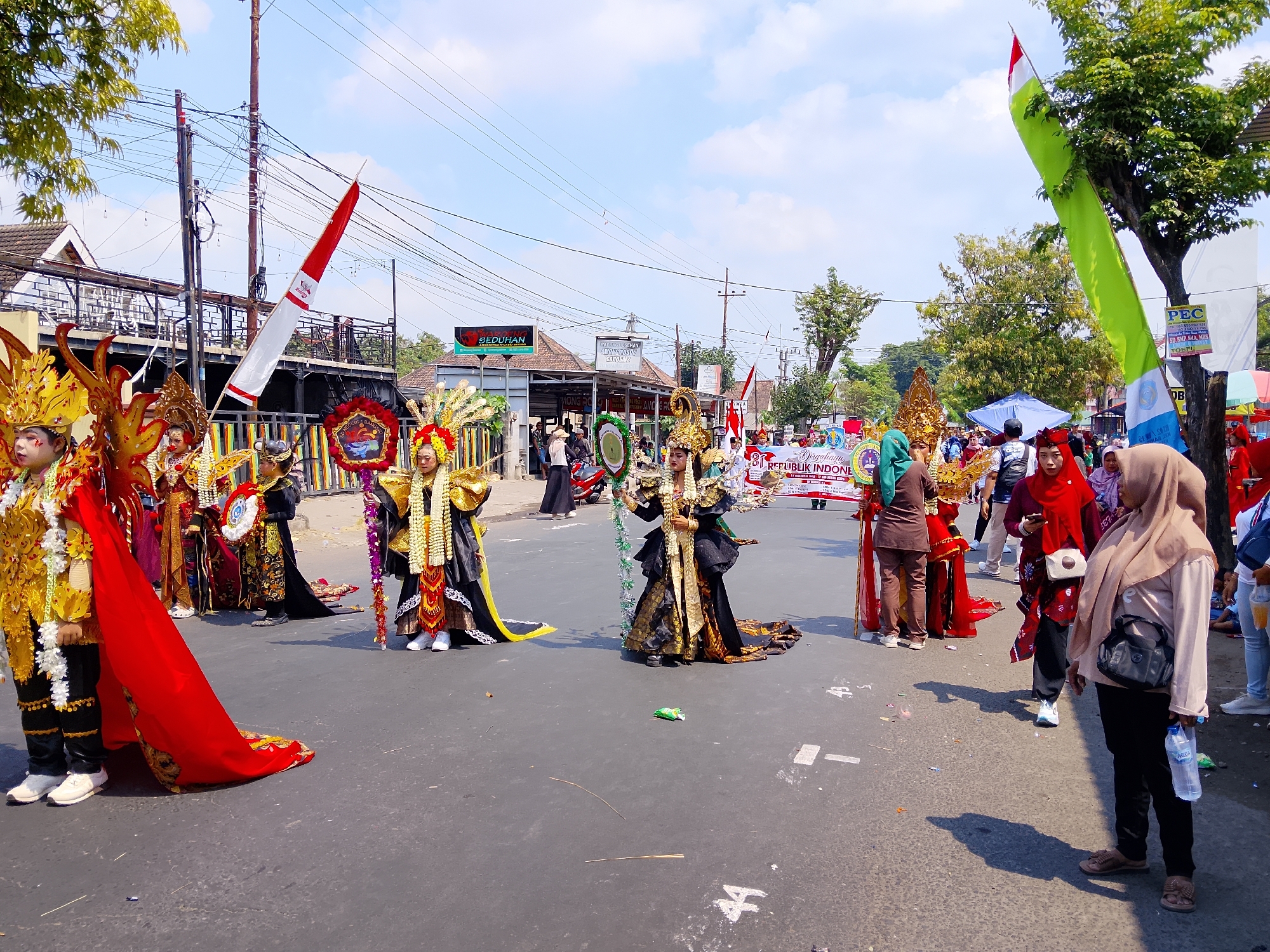 The image depicts a street parade in Kertosono, Indonesia, celebrating the 77th anniversary of Indonesian Independence. Several individuals in elaborate costumes, adorned with gold, red, and black fabrics, are marching in the street. Some wear tall, ornate headpieces with feathers and intricate designs. One person in the foreground on the left is wearing a bright yellow costume with large, red fabric draped over their shoulder. Another individual in the center is dressed in a black and gold elaborate outfit, holding a staff decorated with green and white elements. People in the background, dressed in more casual attire and hijabs, are watching the parade from the sidewalks. Indonesian flags, red and white and green, are visible, along with banners that read "DIRGAHAYU REPUBLIK INDONESIA" (Long Live the Republic of Indonesia). The scene takes place on an asphalt road with buildings and trees lining the sides. The sky is blue with scattered clouds.