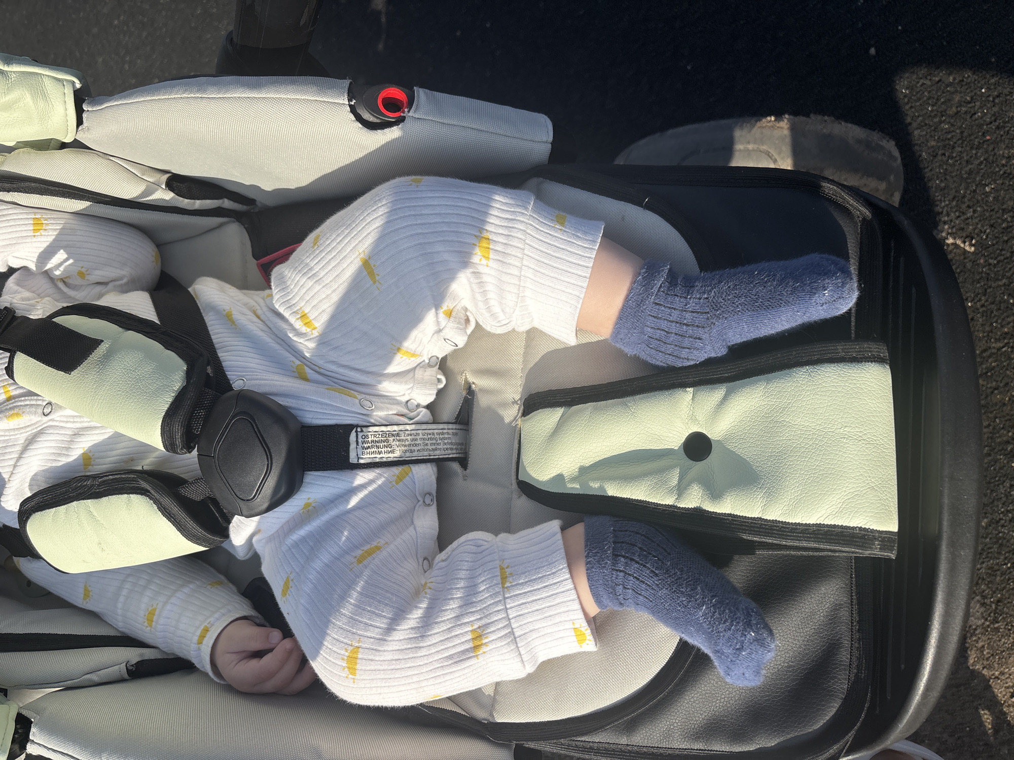 A baby wearing blue socks and a white, long-sleeved onesie with small yellow sun designs is strapped into a stroller. The stroller is light gray with a green padded area where the baby's legs rest. A black harness strap is fastened across the baby's midsection. A warning label is visible on the harness strap. Part of a shoe is visible in the upper right of the image. The stroller is on asphalt. The image was most likely taken outdoors in daylight. The location in the city of Раздолье, Russia cannot be determined from the image.