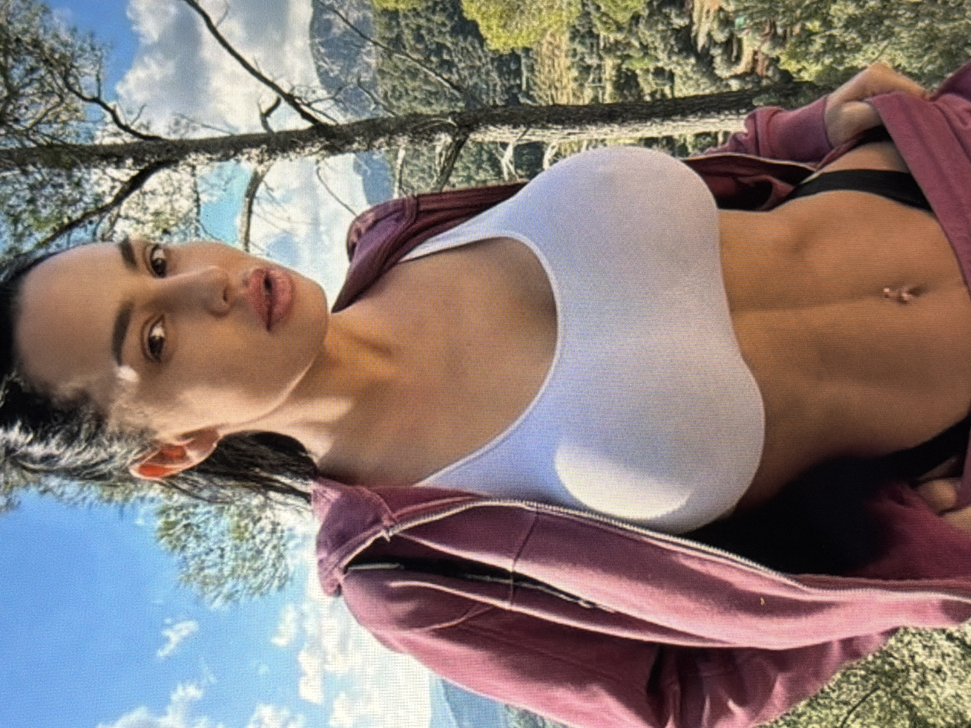 A woman stands outdoors, facing the camera, with a wooded hillside and mountains in the background. She has dark hair pulled back and is wearing a maroon zip-up hoodie over a white sports bra and black bottoms. Her navel is visible, adorned with a piercing. The scene appears to be a natural landscape, potentially a hiking trail or viewpoint, with a clear sky dotted with clouds. The image does not contain any elements that indicate the specific location of Shumen, Bulgaria.