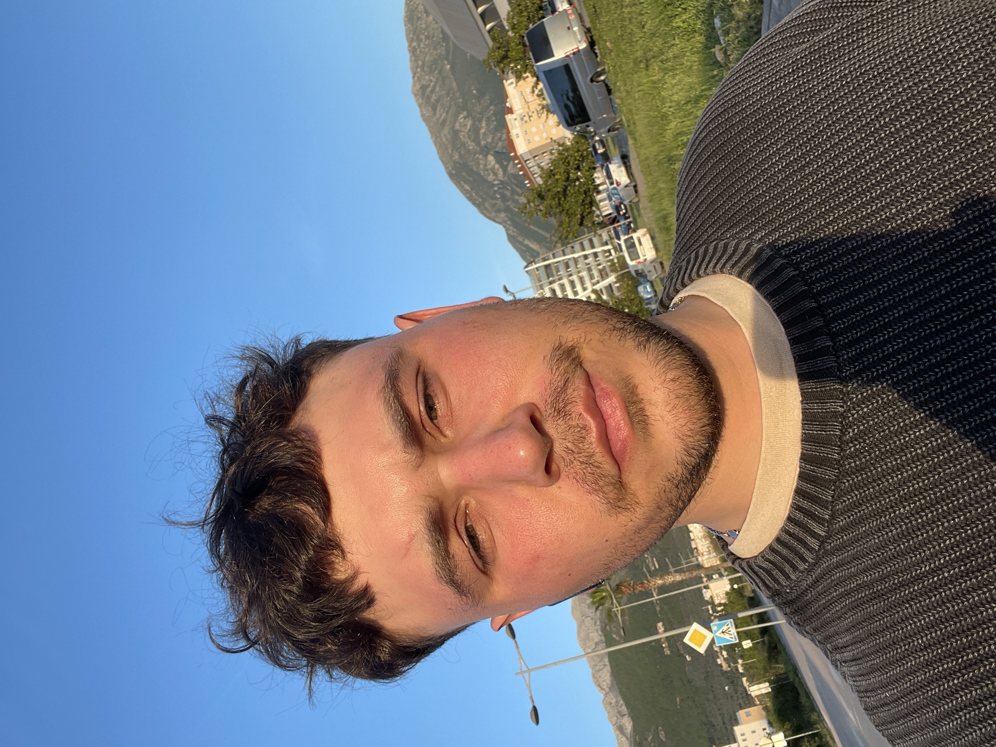 The original image depicts a close-up portrait of a young man, likely taken as a selfie or from a low angle, set against a vibrant outdoor backdrop. The man, possibly in his late teens or twenties, occupies the left side of the frame, looking directly at the camera with a neutral expression, possibly a subtle smile. He has dark, curly hair and a short beard or stubble. His skin appears warm-toned, illuminated brightly by the sun, with some natural redness on his cheeks and forehead. He is wearing a dark grey or charcoal knitted sweater over a light-colored (cream or beige) crew neck undershirt, with a slender silver chain visible around his neck.
The setting is outdoors, under a clear, bright blue sky that dominates the upper left portion of the background. Behind the man, a scenic landscape unfolds, featuring large, rugged mountains in the distance, characteristic of a coastal or mountainous region. In the mid-ground, there are urban or suburban elements including various buildings (some multi-story, possibly residential) and what appears to be a street or road with parked vehicles and possibly moving traffic. Green grass or low vegetation is visible on a slope. The scene suggests late afternoon or early evening, often referred to as the "golden hour," given the warm, bright sunlight creating soft highlights and shadows.
Notable objects in the background include two distinct street signs: a yellow diamond-shaped sign (commonly indicating a priority road in Europe) and a blue square sign with a white upward arrow. Street lights are also visible. The location context of Bar, Montenegro, aligns with the visual cues of mountains, coastal architecture, and bright, sunny weather. There is no legible text visible within the image.