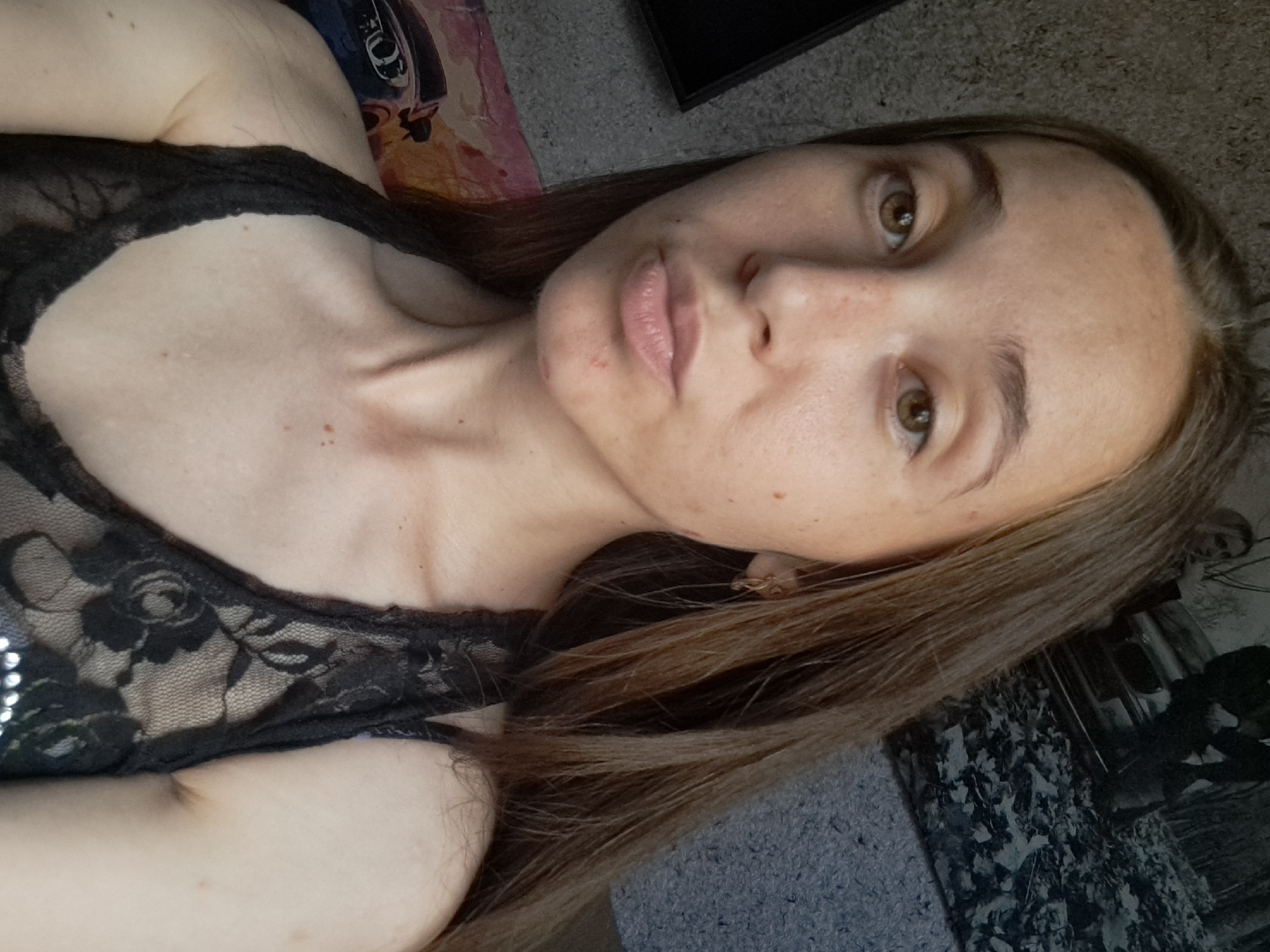 The image shows a close-up, head and shoulders selfie of a young woman with long, brown hair and light brown eyes. She is wearing a black lace top. Her skin is fair with some freckles. She is looking directly at the camera with a neutral expression. The background is out of focus, but appears to be an indoor setting with a textured wall. There are no visible elements that suggest a specific location in Vypasne, Ukraine.