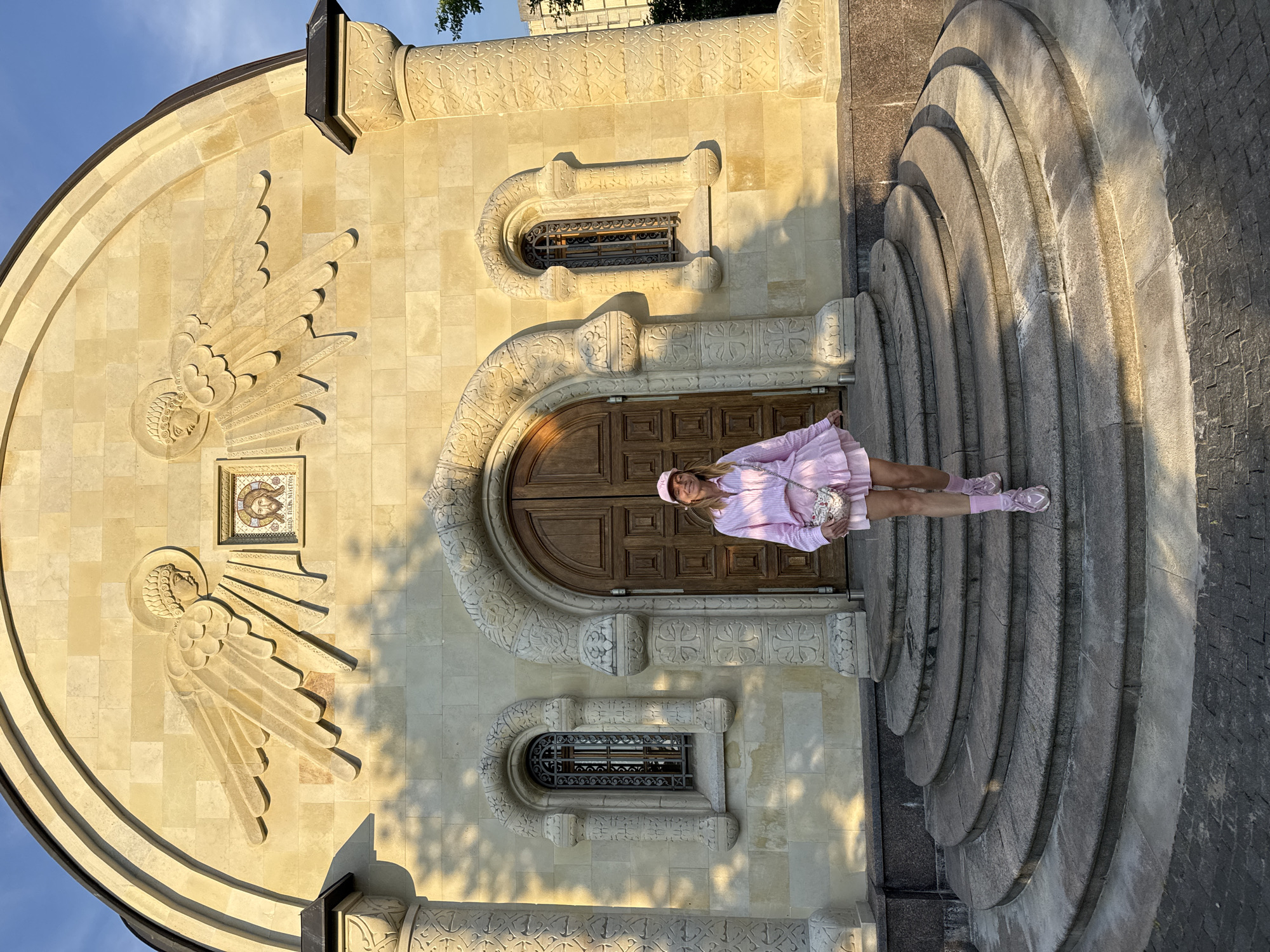 A woman in a pink outfit stands on a curved stone staircase in front of a building in Kirishi, Russia. The building's facade is made of light-colored stone blocks, with decorative carvings around the arched doorway and windows. Above the doorway, there are stone reliefs of angel wings and a mosaic of Jesus' face. The woman is wearing a pink striped dress, a pink cap, and pink sneakers, and is carrying a small, ornate bag. The sun is casting shadows on the facade and staircase, suggesting it is either early morning or late afternoon.