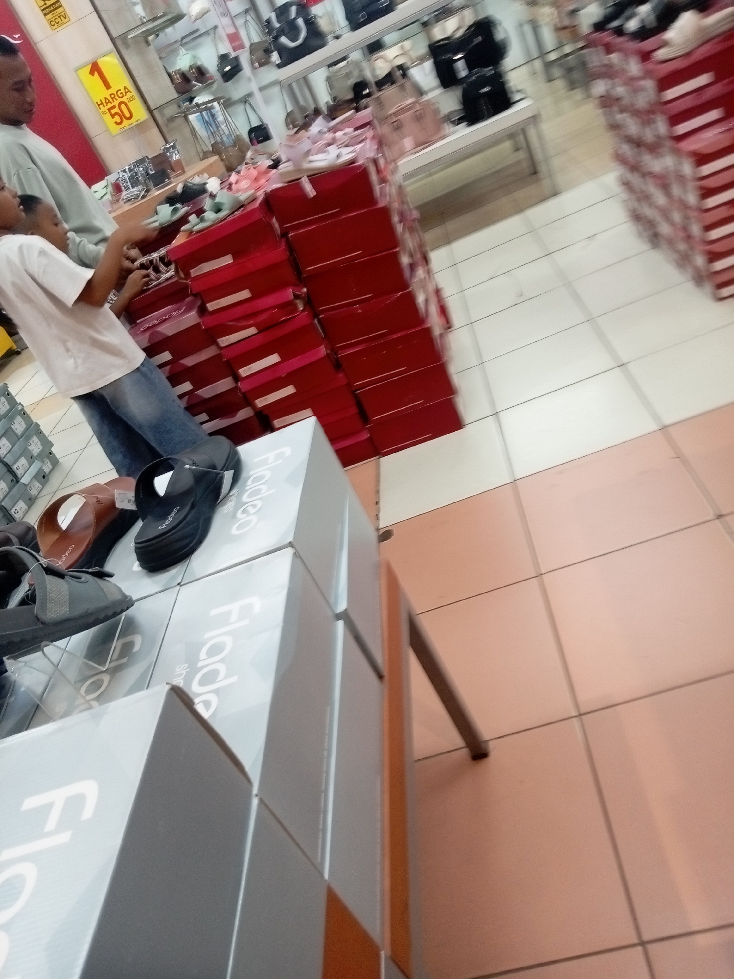 The image captures a retail environment, likely a shoe store in Batam, Indonesia, given the presence of Indonesian text ("HARGA," "PERHATIAN"). In the foreground, several boxes are stacked, displaying branding that appears to read "Flade." On top of these boxes, a pair of black sandals and brown slippers are visible. To the left, a young boy in a white t-shirt and jeans is reaching towards the boxes, while an adult man in a light grey long-sleeved shirt stands behind him, also looking at the merchandise. The background reveals shelves filled with handbags and shoes, and stacks of red boxes, suggesting a well-stocked store. The flooring consists of beige and cream tiles. A yellow sign with "1 HARGA RP. 50.000" is prominently displayed on the left side, indicating a price of 50,000 Indonesian Rupiah.