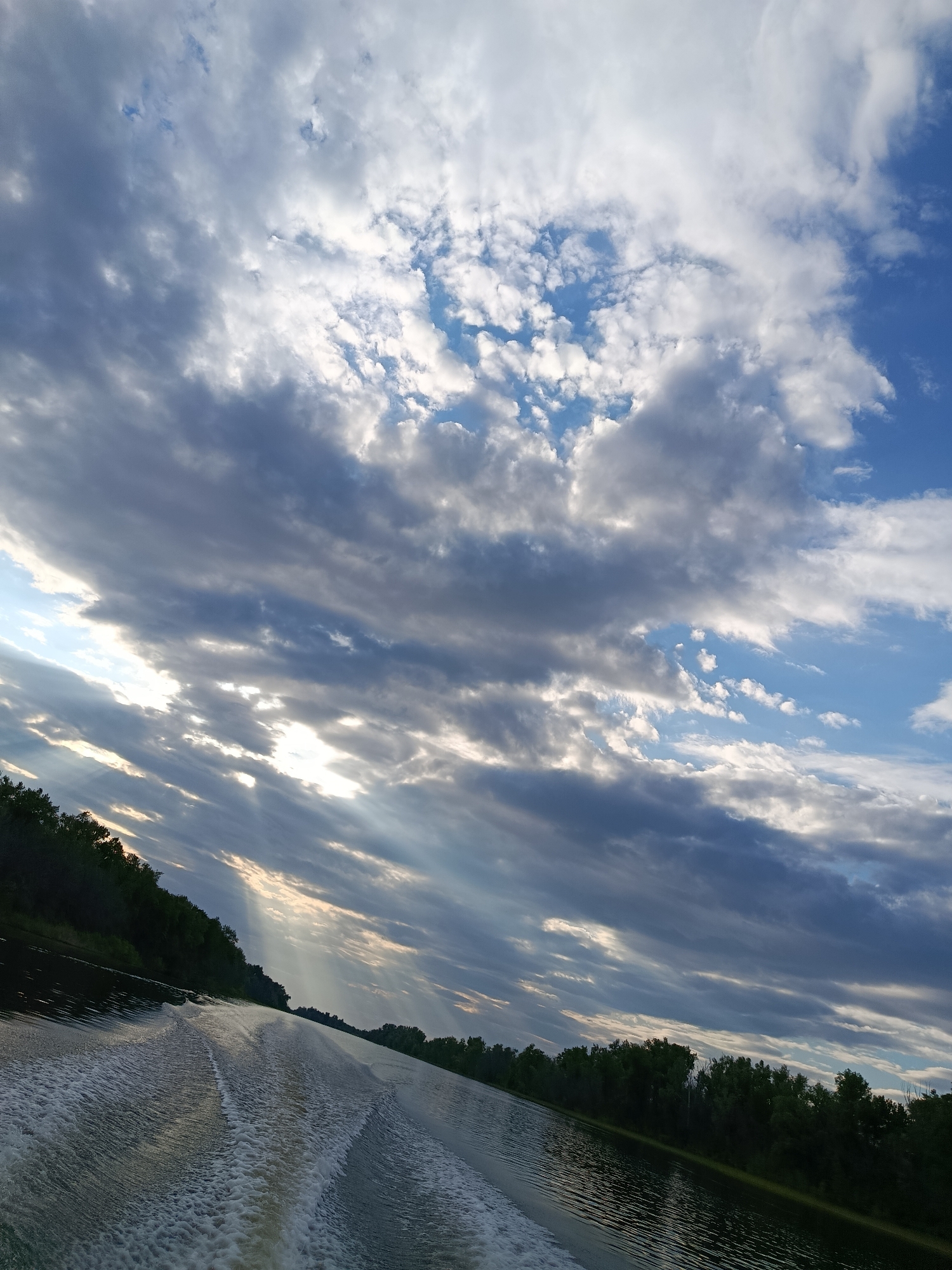 This is a photograph taken from a boat moving on a wide, dark river. The camera is angled to the right, showing the wake of the boat spreading out in foamy white patterns on the water's surface. Along the left bank of the river, a dense line of dark green trees is visible, indicating a wooded shoreline. The sky above is filled with dramatic clouds, a mix of dark, heavy storm clouds and lighter, white cumulus clouds. Sunbeams are breaking through the clouds in several places, casting rays of light downwards. The overall impression is of a dynamic sky over a tranquil natural landscape. The location is identified as Pavlovka, Russia.