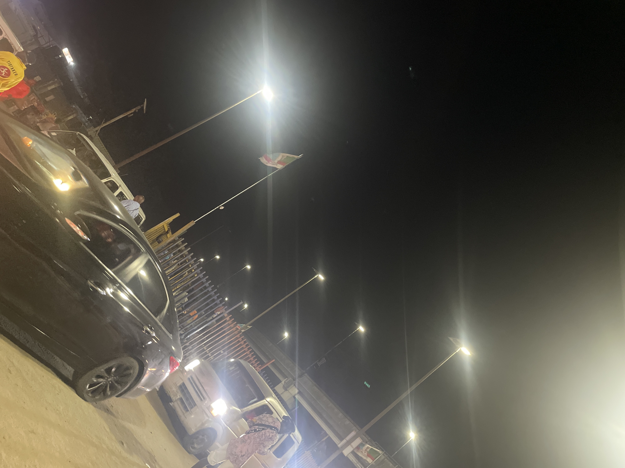 The image shows a night scene in Lekki, Nigeria. A dark-colored car is parked on the left side of the image. There are several other vehicles, including a white van, parked nearby. Streetlights illuminate the scene. A flag is visible atop a pole in the distance. A man in a checkered shirt is standing near one of the vans.
