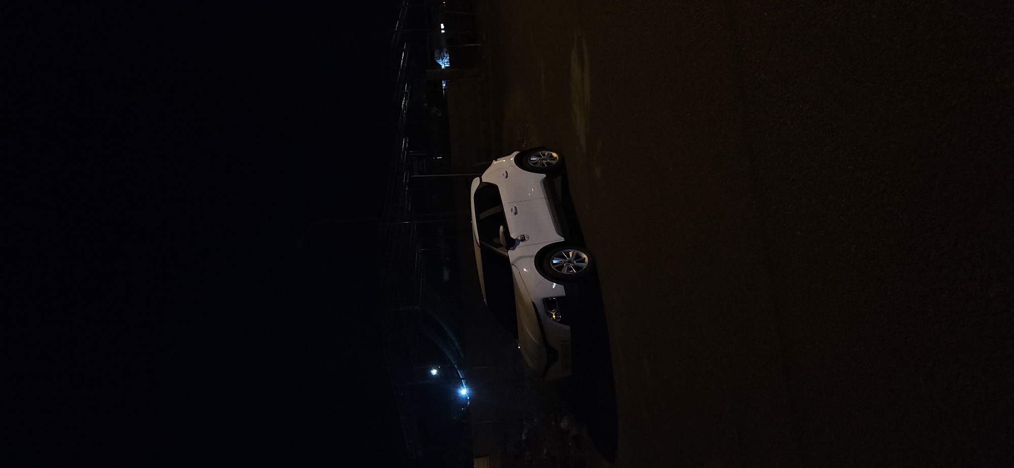 A nighttime scene in Bac Nha Trang Ward, Vietnam, features a white car parked on a dark, textured surface, likely an asphalt road. The image is predominantly dark, with limited visibility due to the nocturnal setting.
The white car, possibly a sedan or hatchback, is positioned with its front or rear wheels and silver-colored rims visible. Its body appears clean and undamaged. The ground immediately surrounding the vehicle shows some reflection or sheen, suggesting it might be wet from recent rain. To the left of the car, a tall, dark, lattice-like structure, resembling scaffolding or a utility tower, is partially visible, extending upwards into the darkness. A prominent blue light source emanates from or near this structure, casting a subtle glow. The sky above is completely dark, indicative of late hours. There are no individuals present, and no active events are captured; the scene portrays a parked vehicle in a still, low-light environment. No text is visible within the frame.