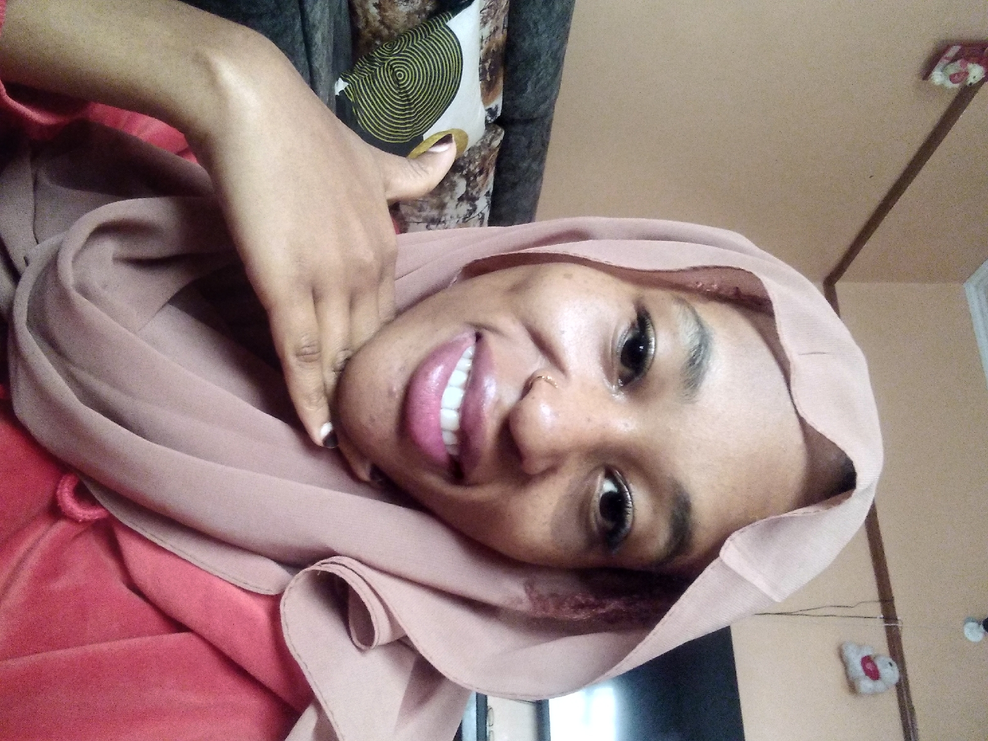 A young woman wearing a pink hijab and a red top is taking a selfie. She is indoors, and behind her, a couch and pillows are visible. On the wall behind her, there are two small decorative items. The lighting suggests it is daytime, likely in a residential setting within the city of Nyeri, Kenya.