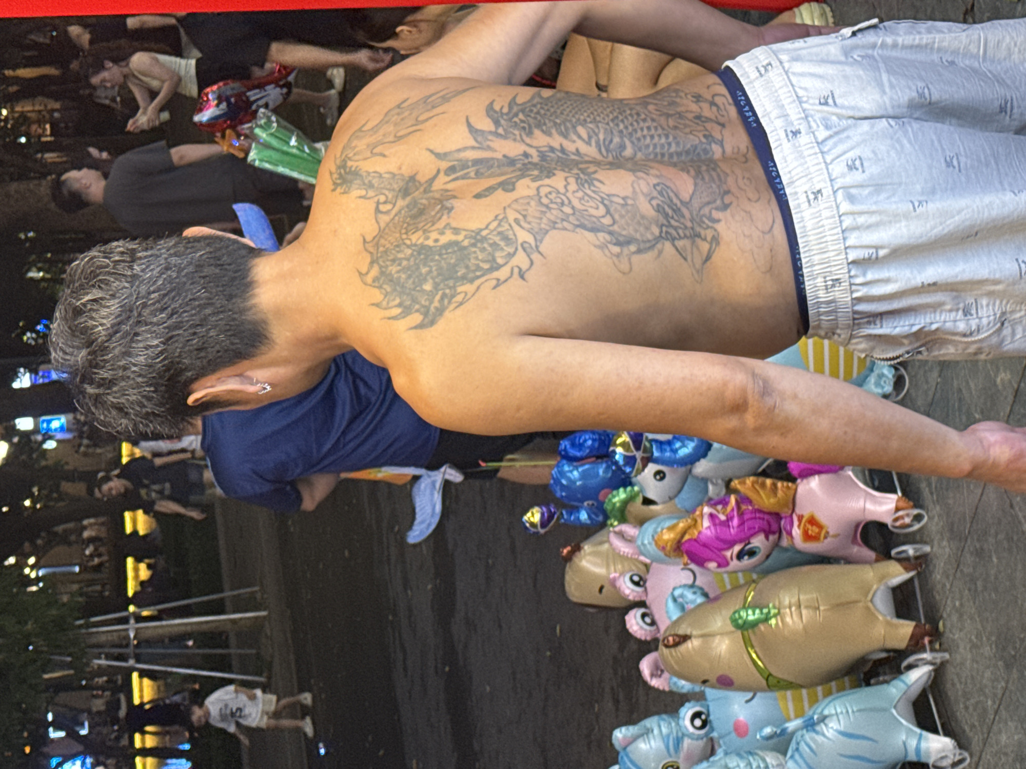 A shirtless man with a large dragon tattoo on his back stands in a public area in Hà Nội, Vietnam, surrounded by inflated cartoon animal balloons. He has short, graying hair and is wearing light-colored shorts. The balloons depict various characters, including cartoon animals with large eyes. In the background, other people can be seen, including a child in a white shirt and shorts. Street lights illuminate the scene.