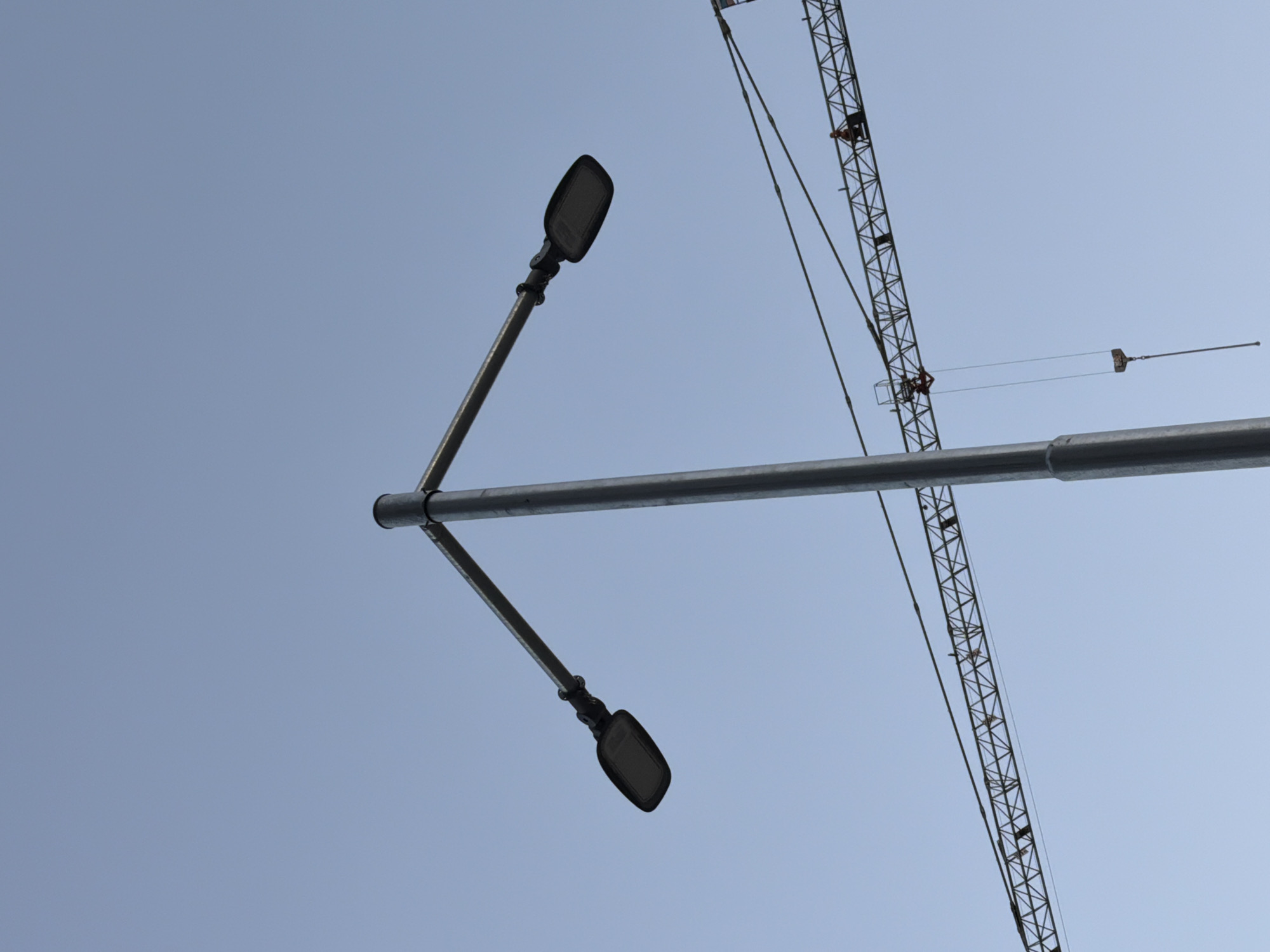 A street lamp with two lights is positioned against a clear, pale blue sky. In the background, a construction crane is visible, with a worker standing on its horizontal arm. The image appears to have been taken during the daytime. The scene is generic and does not contain specific landmarks or indicators to pinpoint a location within the city of Plovdiv, Bulgaria.