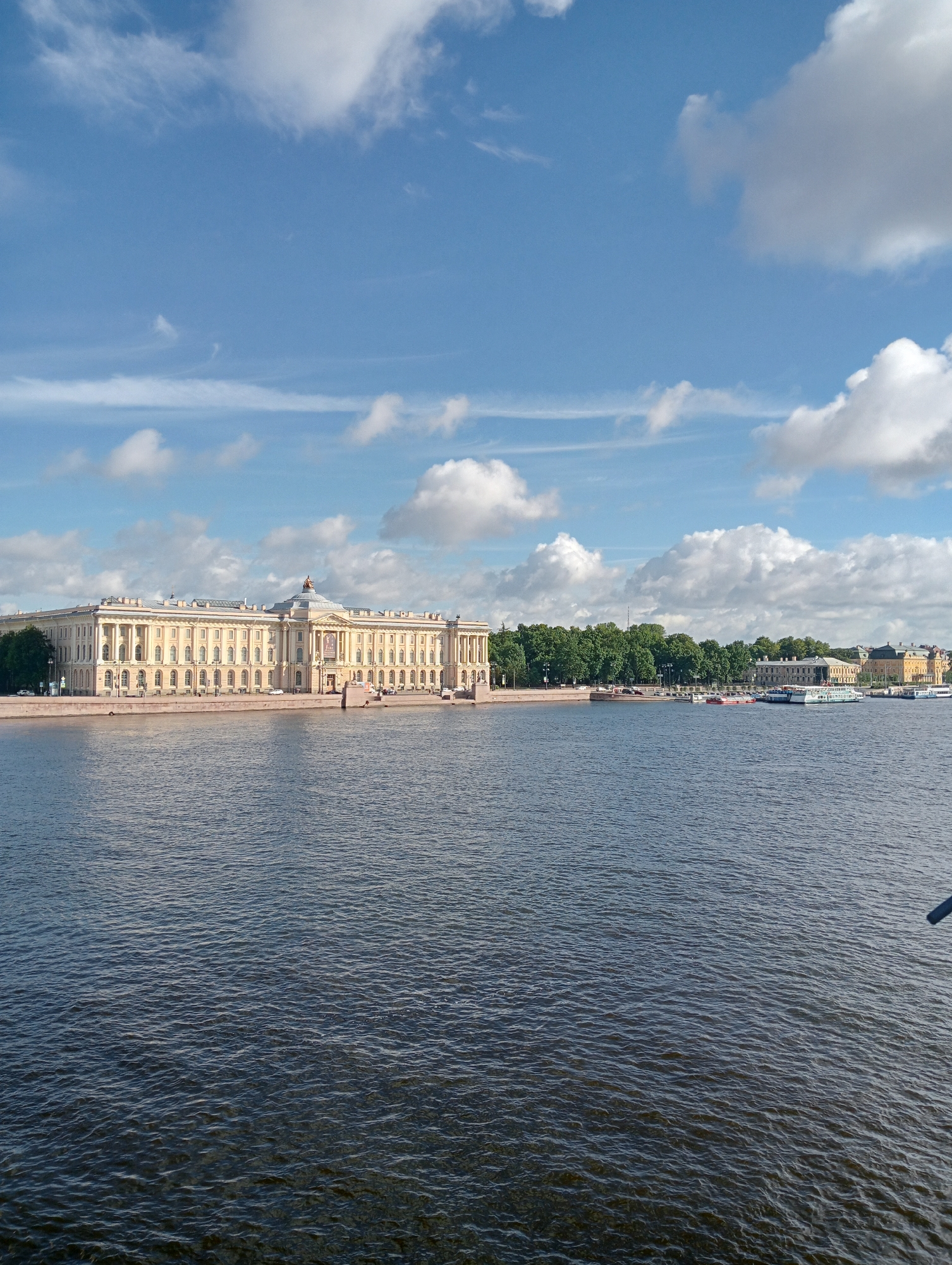 A long, yellow, neoclassical building with a prominent dome and many columns stretches across the middle ground of the image, situated on the bank of a wide, dark body of water. The building appears to be the Kunstkamera. The water in the foreground is choppy with small waves. Behind the building, a line of dense green trees separates it from a hazy cityscape with yellow and white buildings. Several boats are visible on the water in front of the treeline, including white cruise ships and smaller red vessels. The sky above is a bright blue, dotted with fluffy white clouds. The overall scene suggests a sunny day in Saint Petersburg, Russia, likely along the Neva River.