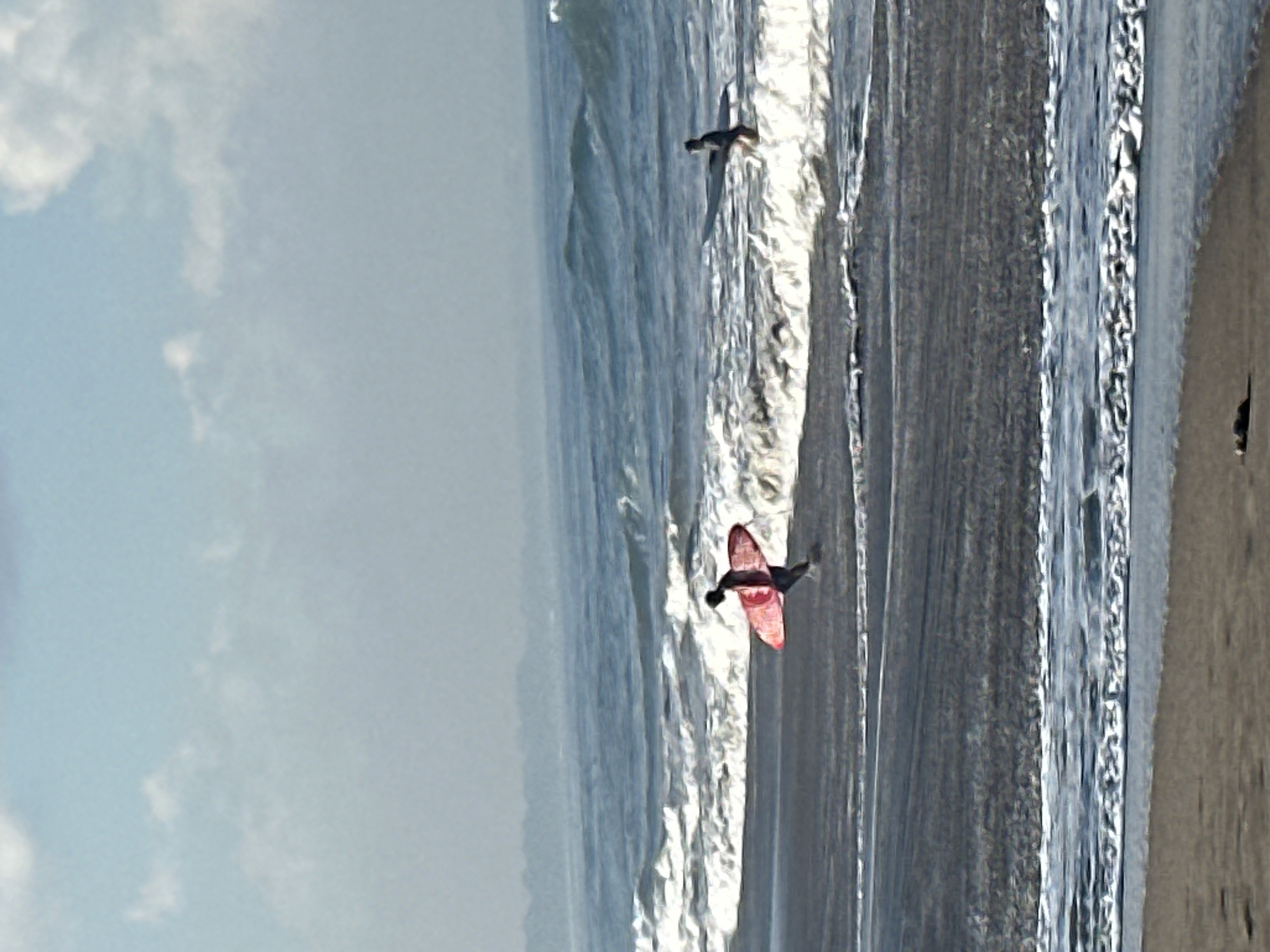 Two individuals are on a dark sand beach in North Kuta, Indonesia, with the ocean in the background. The individual on the left is a surfer, running with a red surfboard held under their right arm. The surfer's body is angled to the right, and they are wearing dark clothing. The individual on the right is further in the water, also appearing to be a surfer, with a light-colored surfboard. They are in a crouched position, facing towards the left. The ocean has moderate waves breaking near the shore. The sky is mostly clear with some scattered white clouds. The overall scene depicts a typical day for surfing activities.