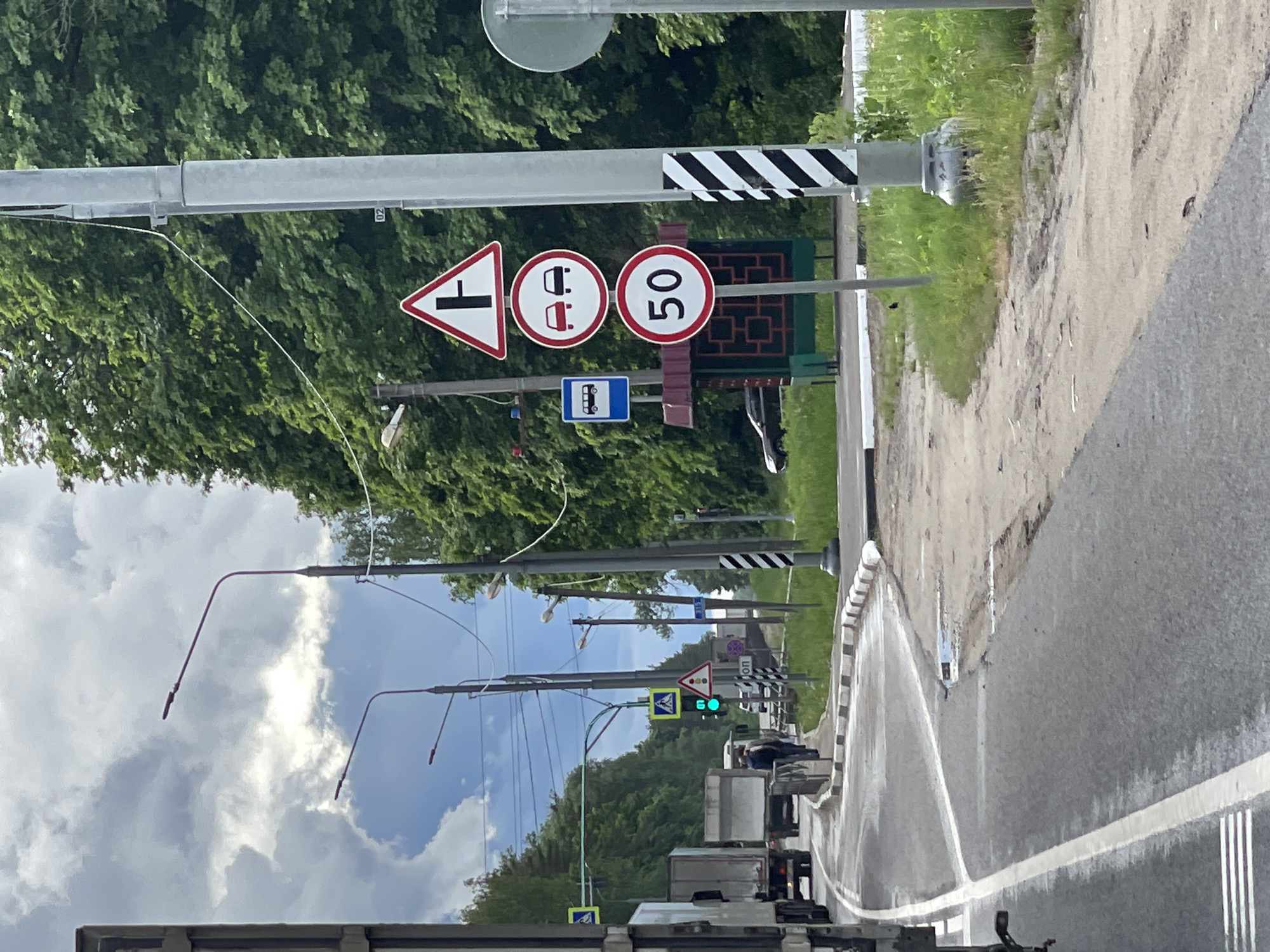 The image shows a road in Ivanovskoye, Russia with several traffic signs. These include a T-intersection warning sign, a sign prohibiting the use of vehicles with trailers, a speed limit sign indicating 50 km/h, and a bus stop sign. Several street lights are visible along the road, with one displaying a green traffic light. Cars and trucks are present on the road. The sky is cloudy. The environment includes trees and greenery alongside the road. The road surface is wet.