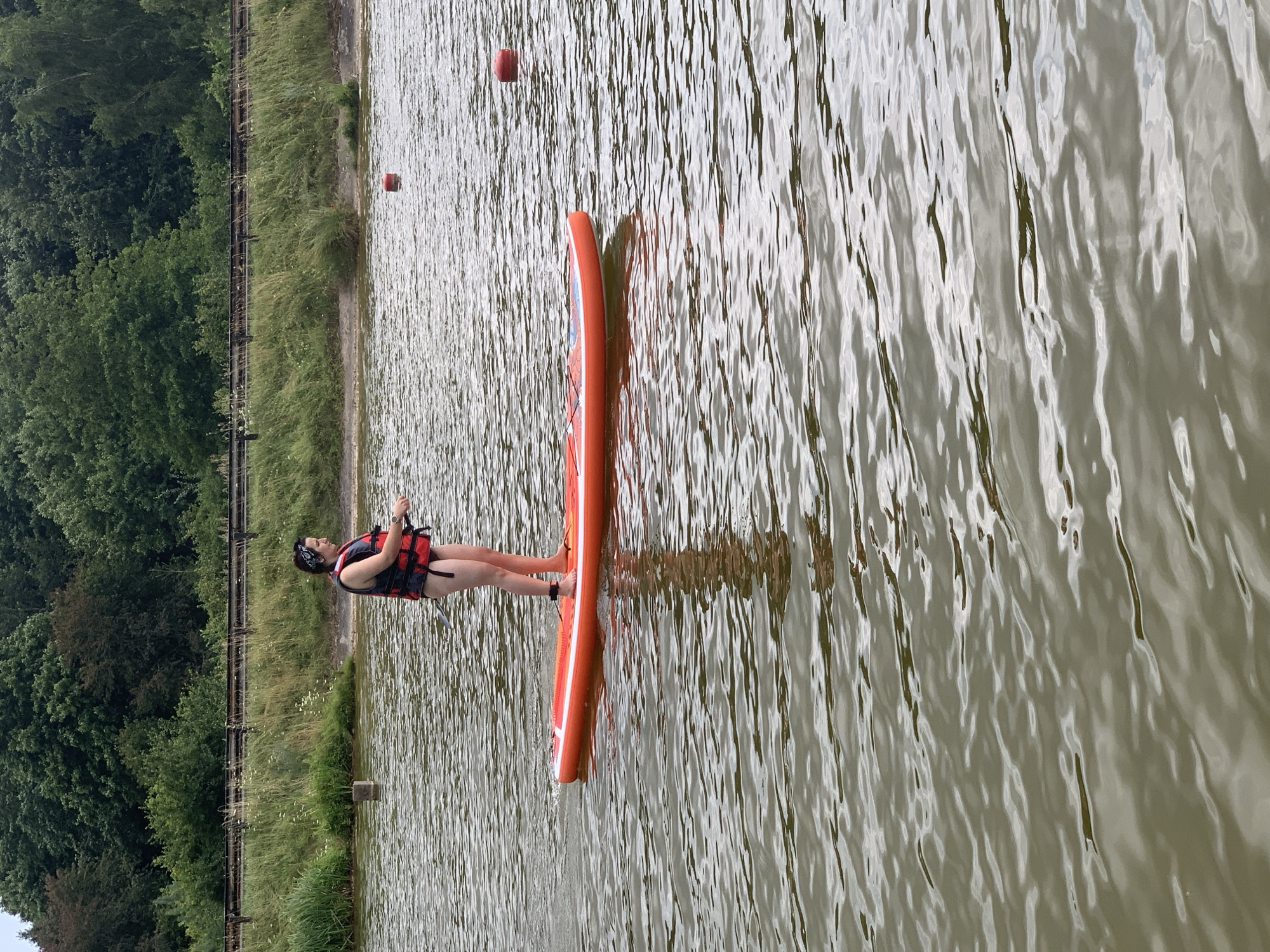 A person stands on an orange paddleboard in a body of water. The person is wearing a red life vest over dark swimwear and a black and white bandana on their head. They are holding a paddle. The paddleboard is floating on the water, which appears murky and has ripples. In the background, there is a shoreline with tall grass and a dense line of trees. There are also two red buoys visible in the water. The scene is outdoors, with overcast lighting.