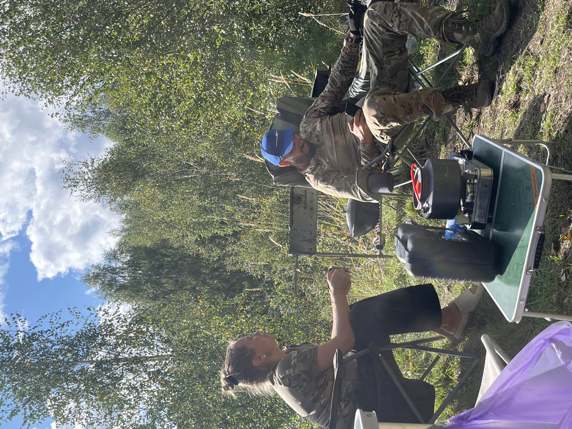 Two people are sitting in camping chairs in a grassy, wooded area. The woman on the left is wearing camouflage clothing and has her hair in a bun. She is holding a mug and looking to her right. The man on the right is wearing a camouflage shirt, pants, and a blue baseball cap. He is looking down at his phone, which he is holding in his hands. In the foreground, there is a small green folding table with a portable stove and a kettle on it. There is also a black case and a purple bag. The background is filled with trees and foliage, and the sky is blue with some white clouds. The scene appears to be in the city of Novyi, Russia, given the context.