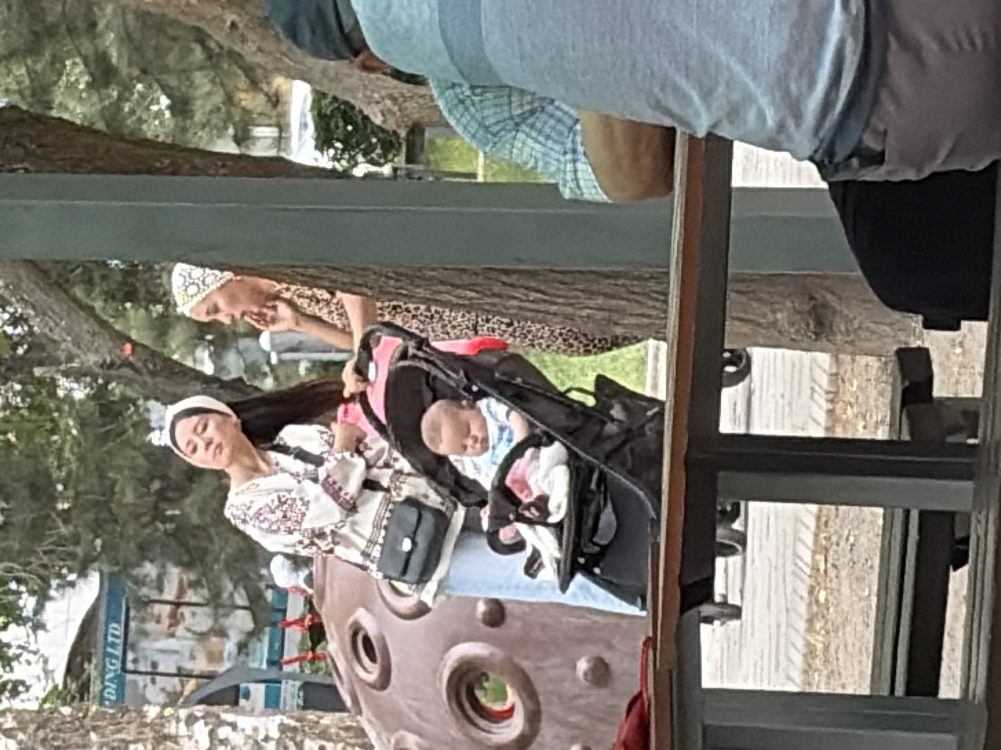 A baby sits in a stroller next to two women. The woman on the left, wearing a white top with embroidered designs and a white headscarf, looks upwards. The woman on the right, in a leopard print top and a white headscarf, touches her face. The baby appears to be wearing a blue and white outfit. In the background, there is a large, circular, textured object that resembles a planet. A person wearing a blue and white plaid shirt is seated at a table in the foreground. The scene appears to be outdoors, with trees and a building visible.